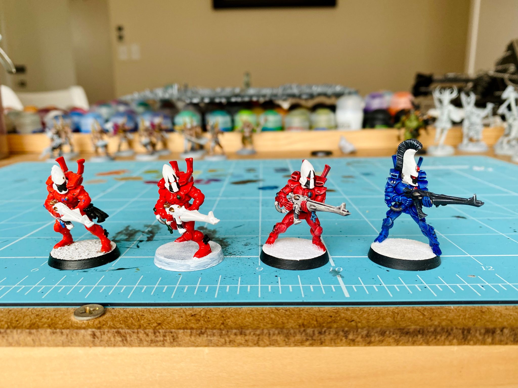 A photo of three Eldar Guardian miniatures and a Dire Avenger. The Guardians are painted red with white helmets, the left-most one has no washes or dry-brushing and is very plain. The next is richer red but still no proper washes or drybrushing. Next is one I improved by adding Contrast paint and a drybrush, and the Dire Avenger is painted rich dark blue with nice drybrushing and highlights.