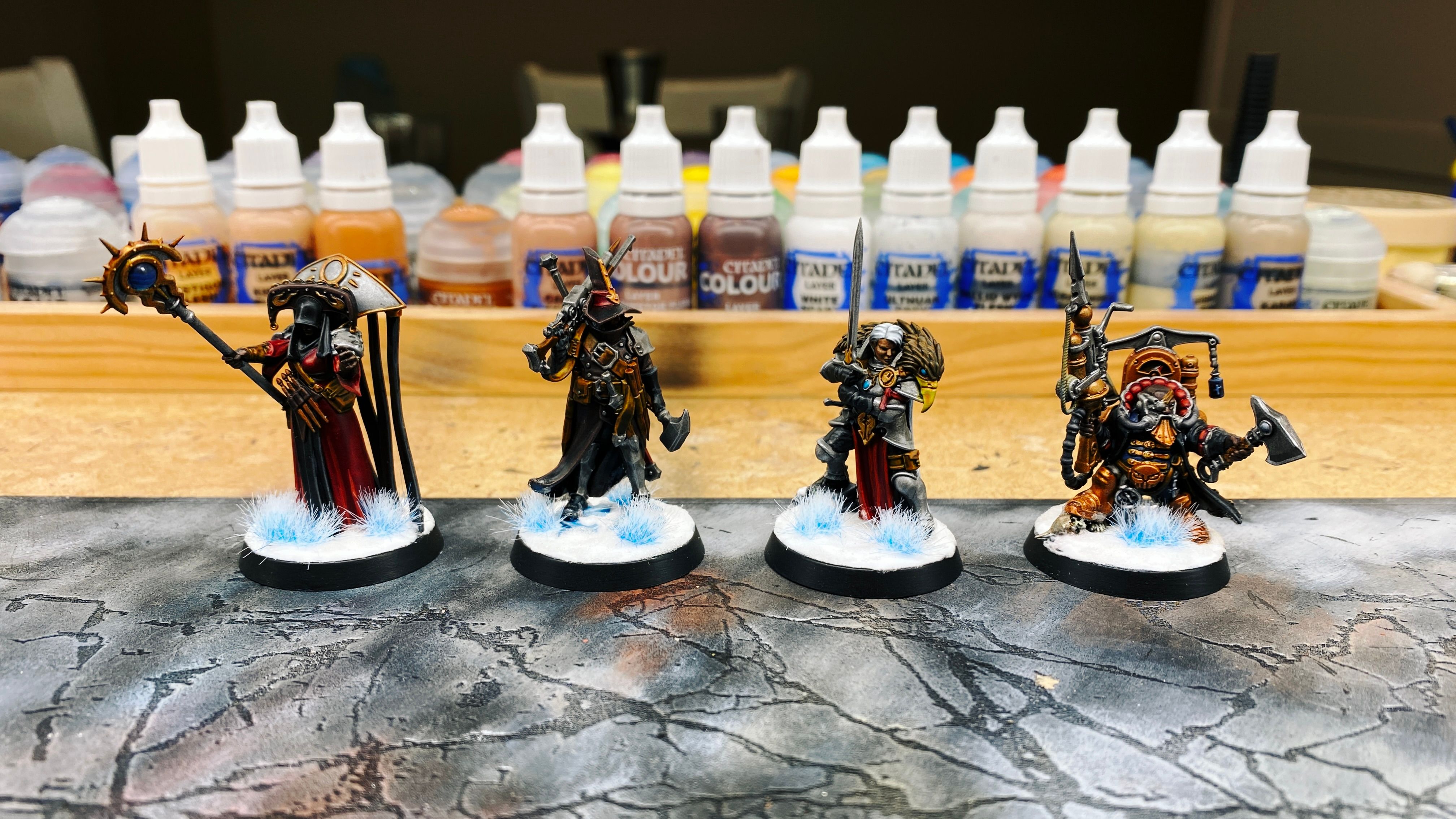 A photo of four painted miniatures:
- A vampire hunter with the standard pointy hat and cool dark brown leather cloak holding a shotgun-looking thing, and with many wooden stakes about his person.
- A heavily-armoured warrior woman with white hair holding a large sword in front of her, with the head of a gryphon draped over her shoulders.
- A dwarf with steampunk-looking armour holding an axe and a steampunk-looking gun.
- A priest in long deep rich red robes with a black hood on and a big elaborate, uh... metallic thing on her back that a bunch of strips of fabric are hanging from. She holding a staff and has her hand out like she's about to cast a spell.

The bases are all painted to look like snow, complete with sparkles, and have icy blue grass tufts on them.