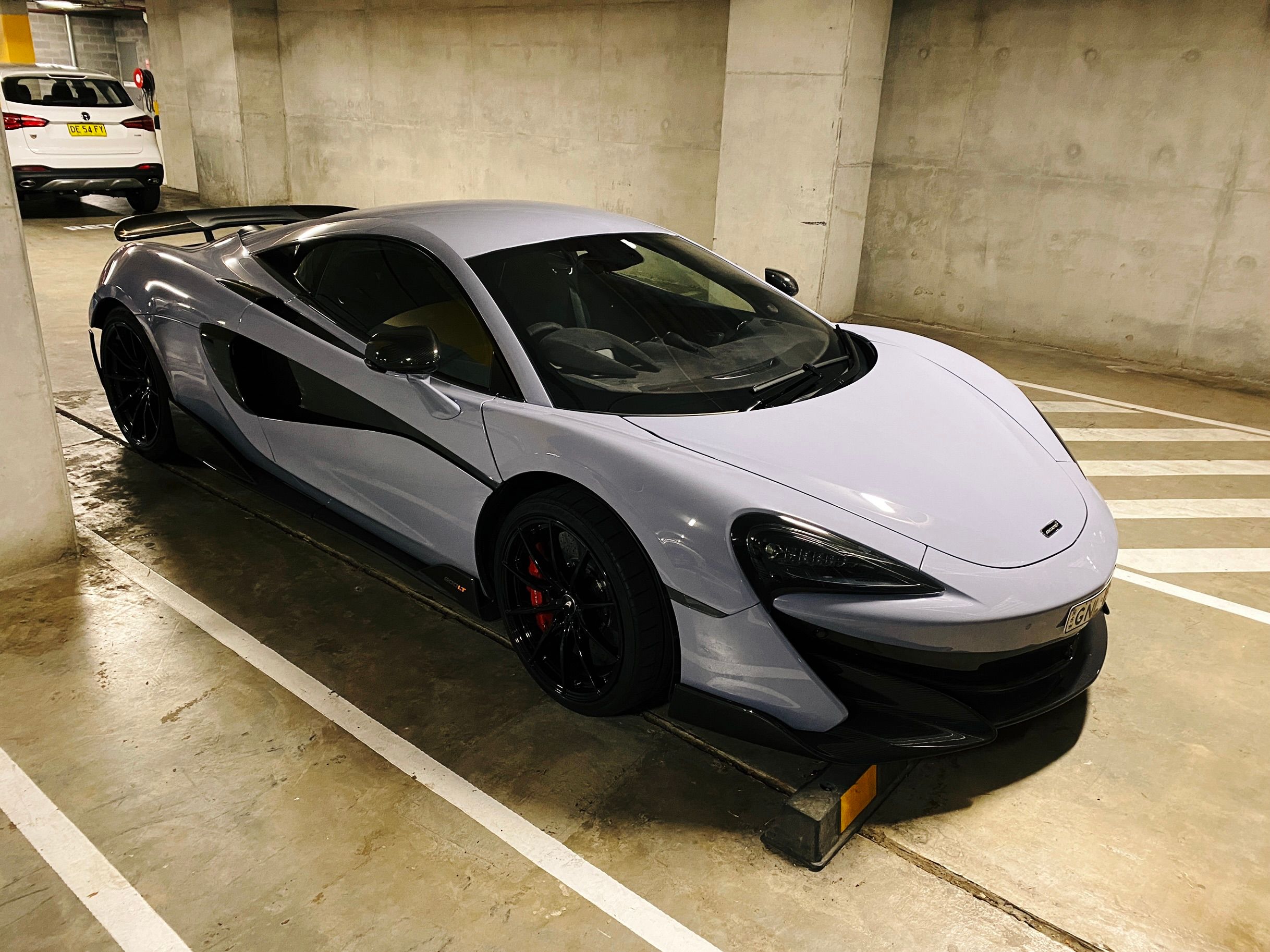 A photo of a pale bluey/purpley McLaren supercar parked in a normal-looking concrete underground carpark. It's very low and wide, and is extremely aerodynamic-looking.