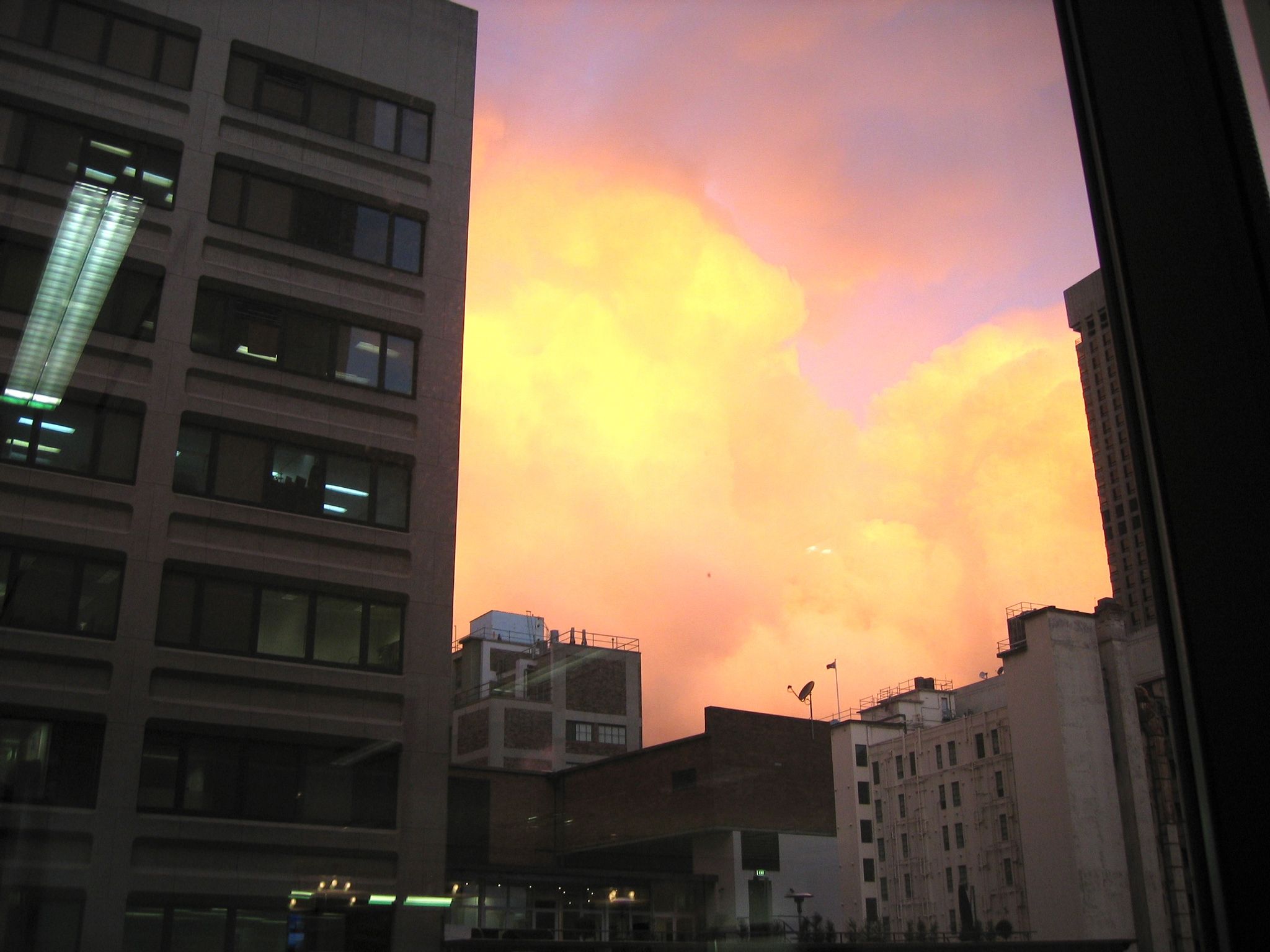 A photo looking out the window of my work desk with very fluffy, very orange clouds in between two tall buildings.