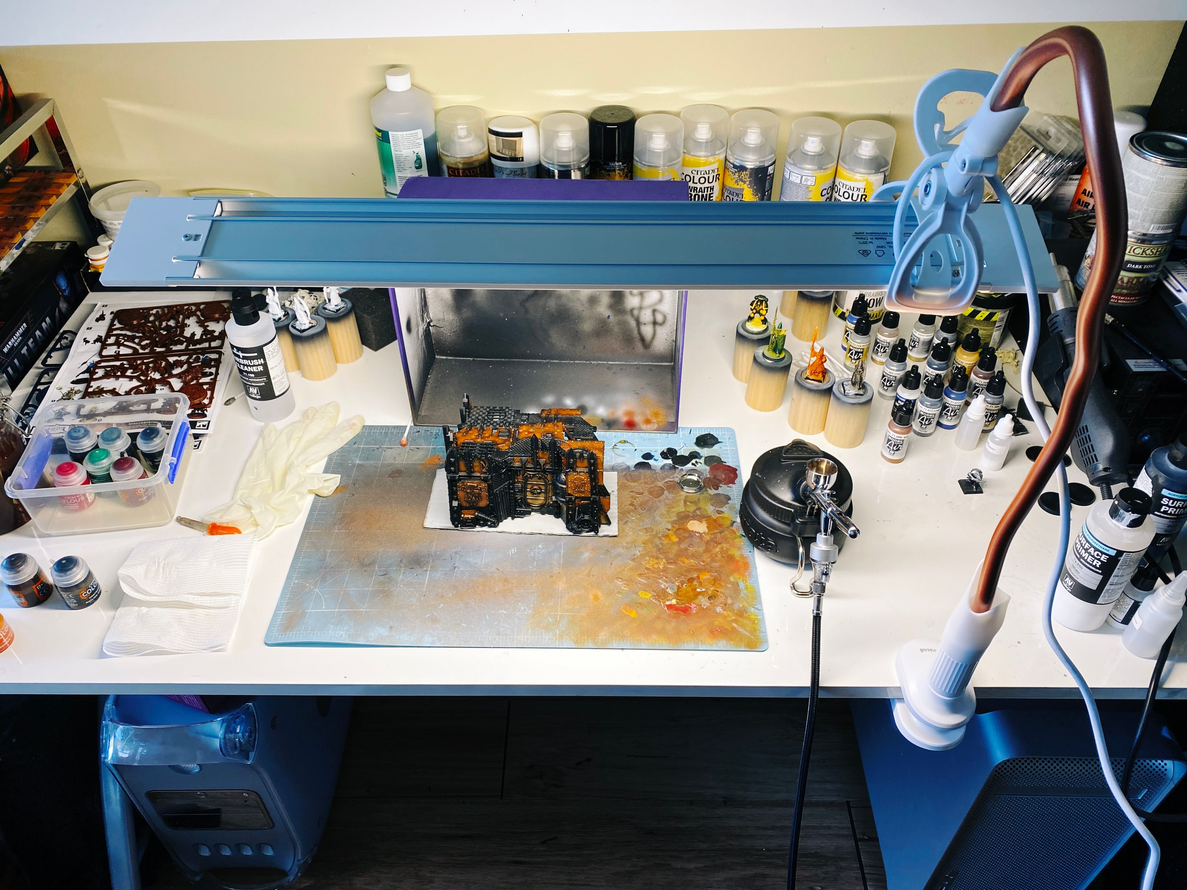 A photo of a very messy desk with an airbrush sitting on it, a piece of junkyard-looking terrain from Warhammer 40,000, all of which is being brightly lit from above by an long narrow LED light that's pointing downwards. The light is being held in place by a gooseneck phone holder that's attached to the side of the desk.