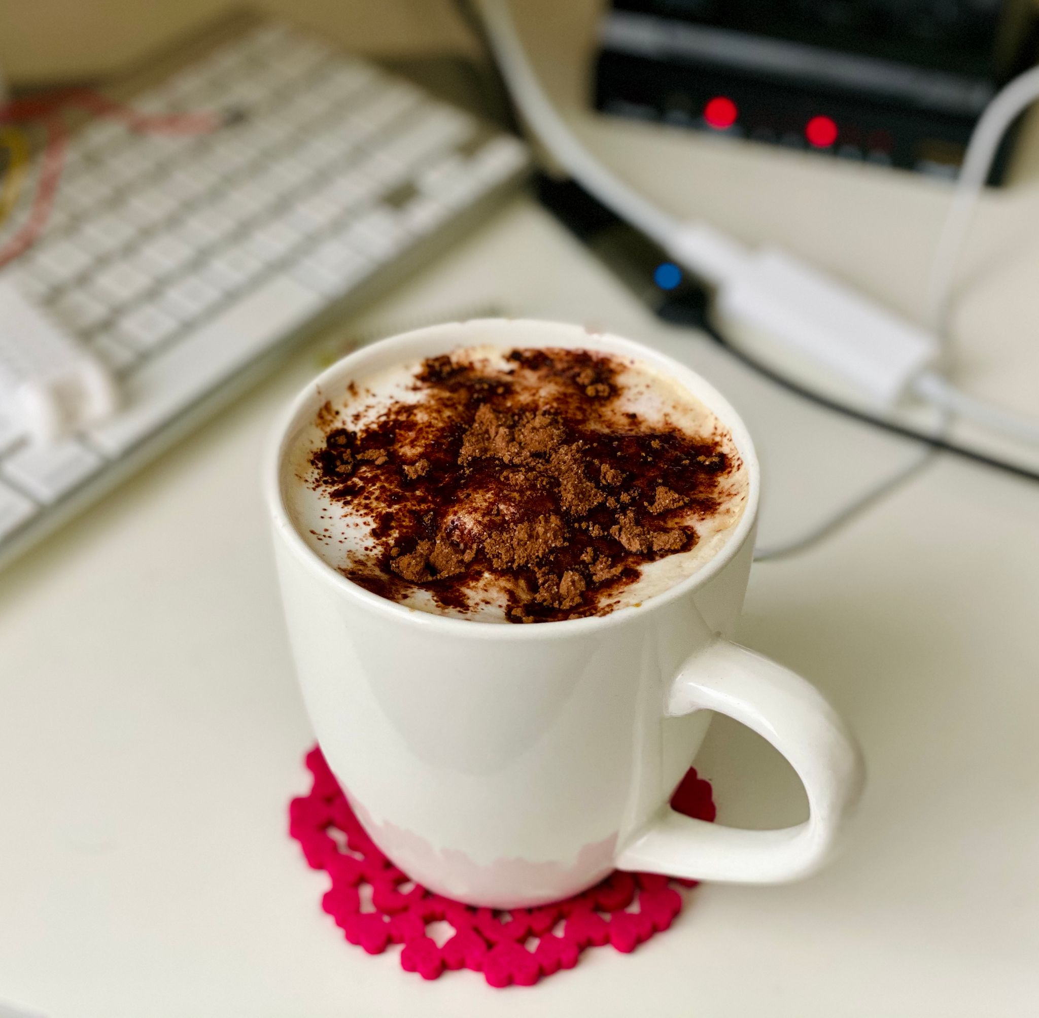 A photo of a mug of coffee, ostensibly a flat white but it has dark chocolate hot chocolate mix sprinkled on top of the milk foam.