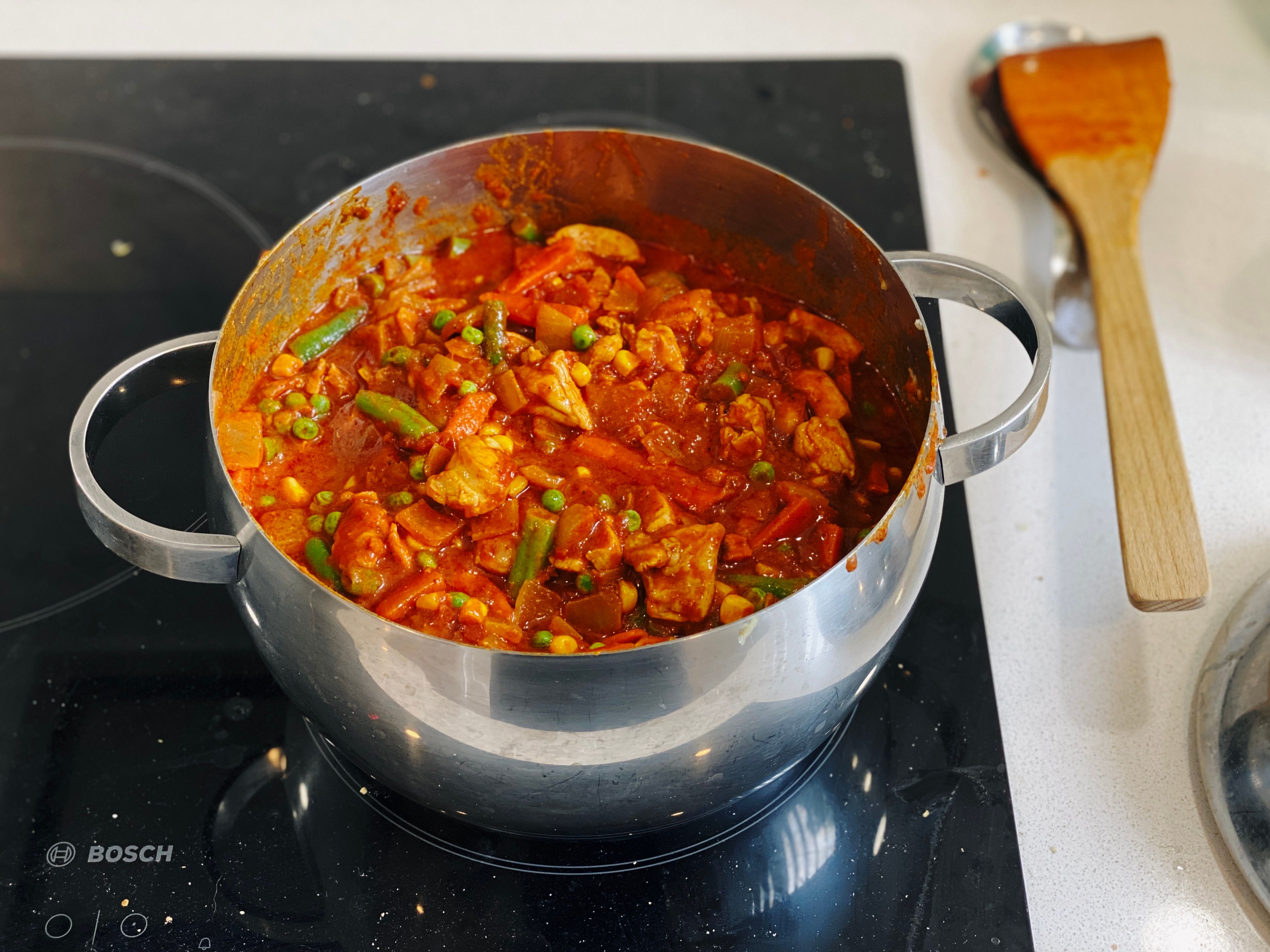 A big saucepan with a rich red chicken curry cooking in it. There's green beans, corns, carrot, and peas visible as well.