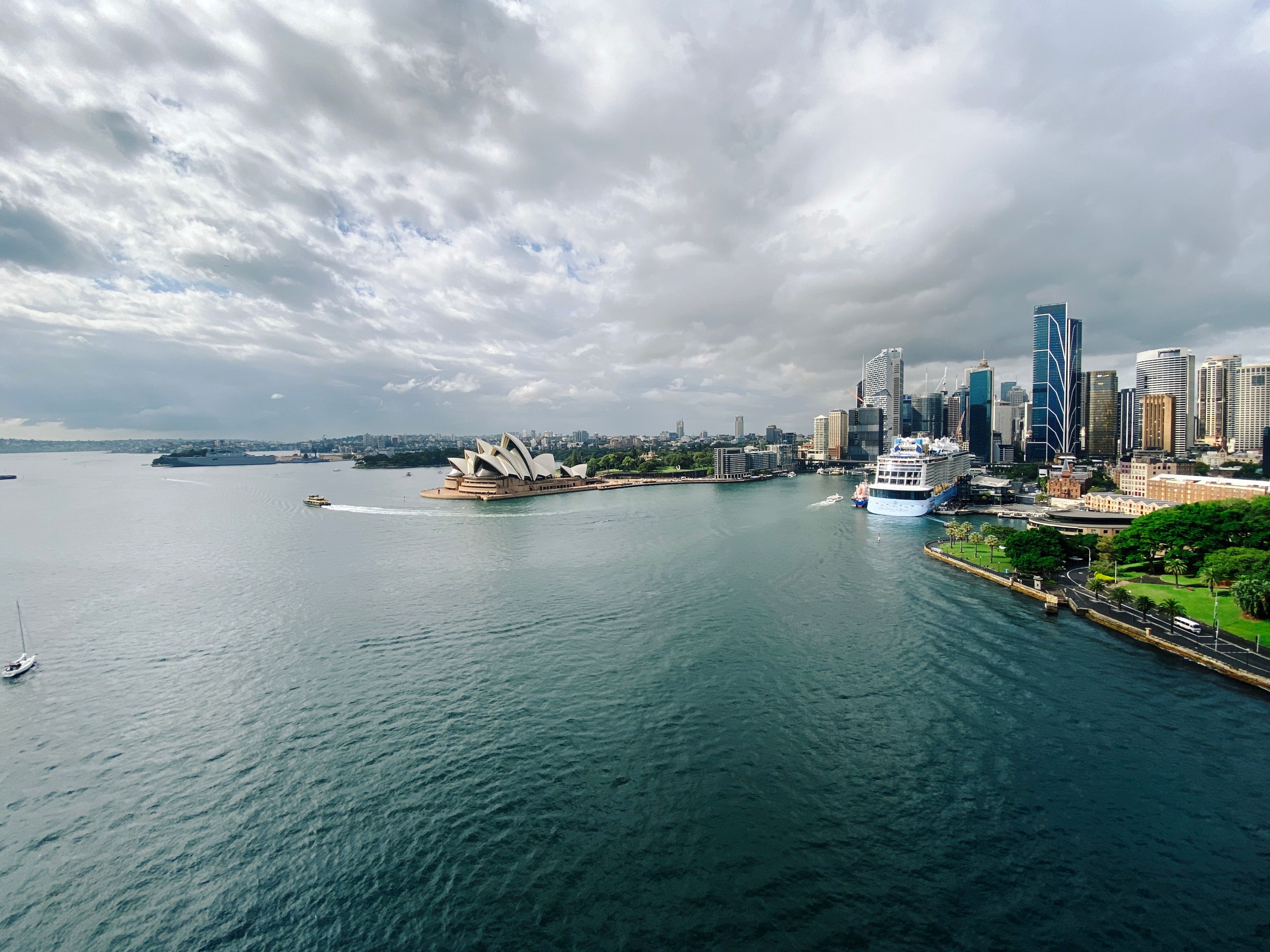 A wide-angle view taken from the middle of the Sydney Harbour Bridge looking over at the Sydney Opera House. There's a GIANT cruise ship moored at Circular Quay to the right, behind which are a bunch of tall buildings. The sky is all grey and cloudy with very small patches of blue poking through.