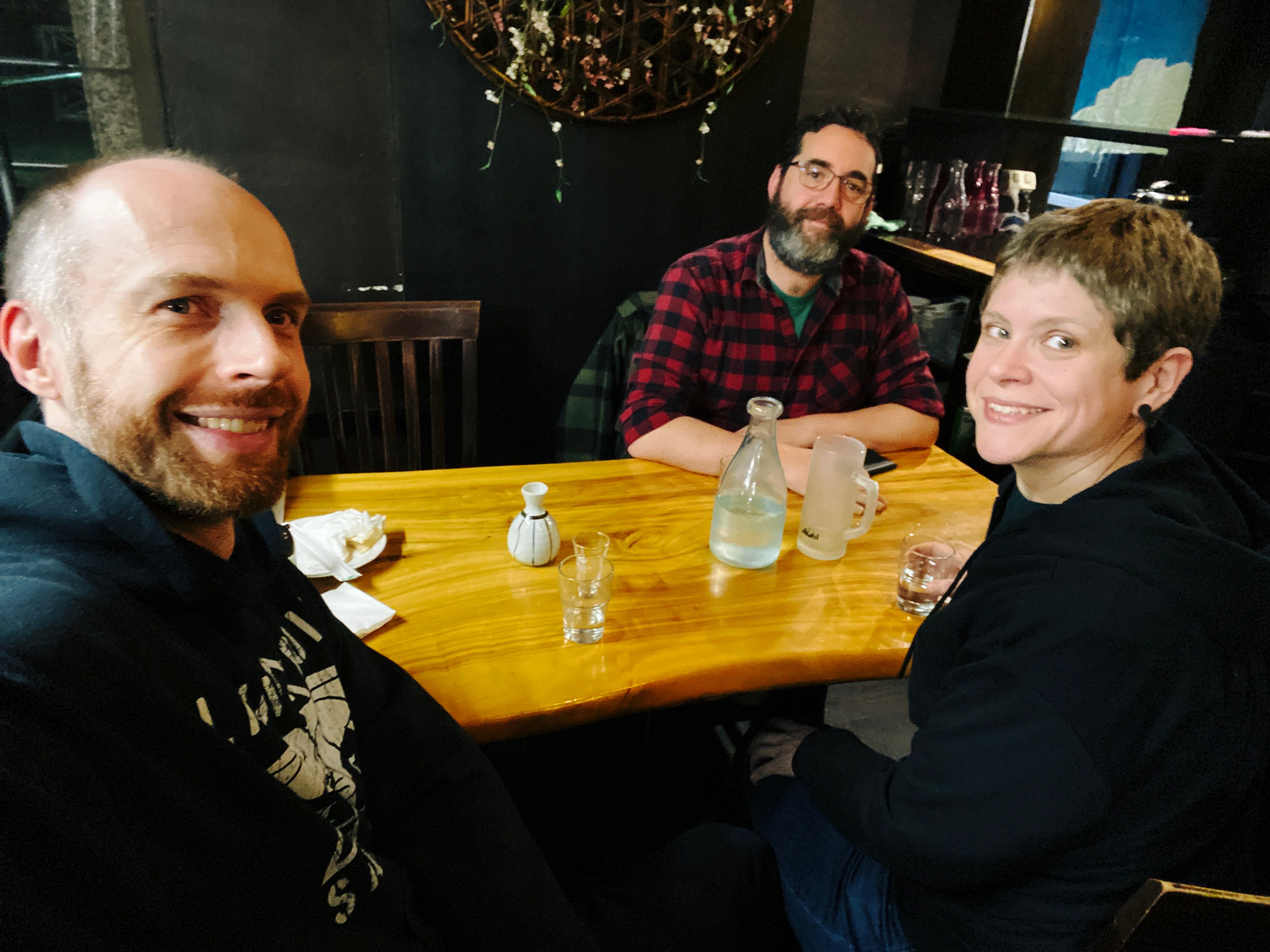 A selfie of three people sitting around a dining table. There's me, a white man with a red beard and short brown hair, Pete, a white man with slightly longer brown hair and a much larger brown beard, and Kristina, a white woman with short brown hair. We're all smiling at the camera.