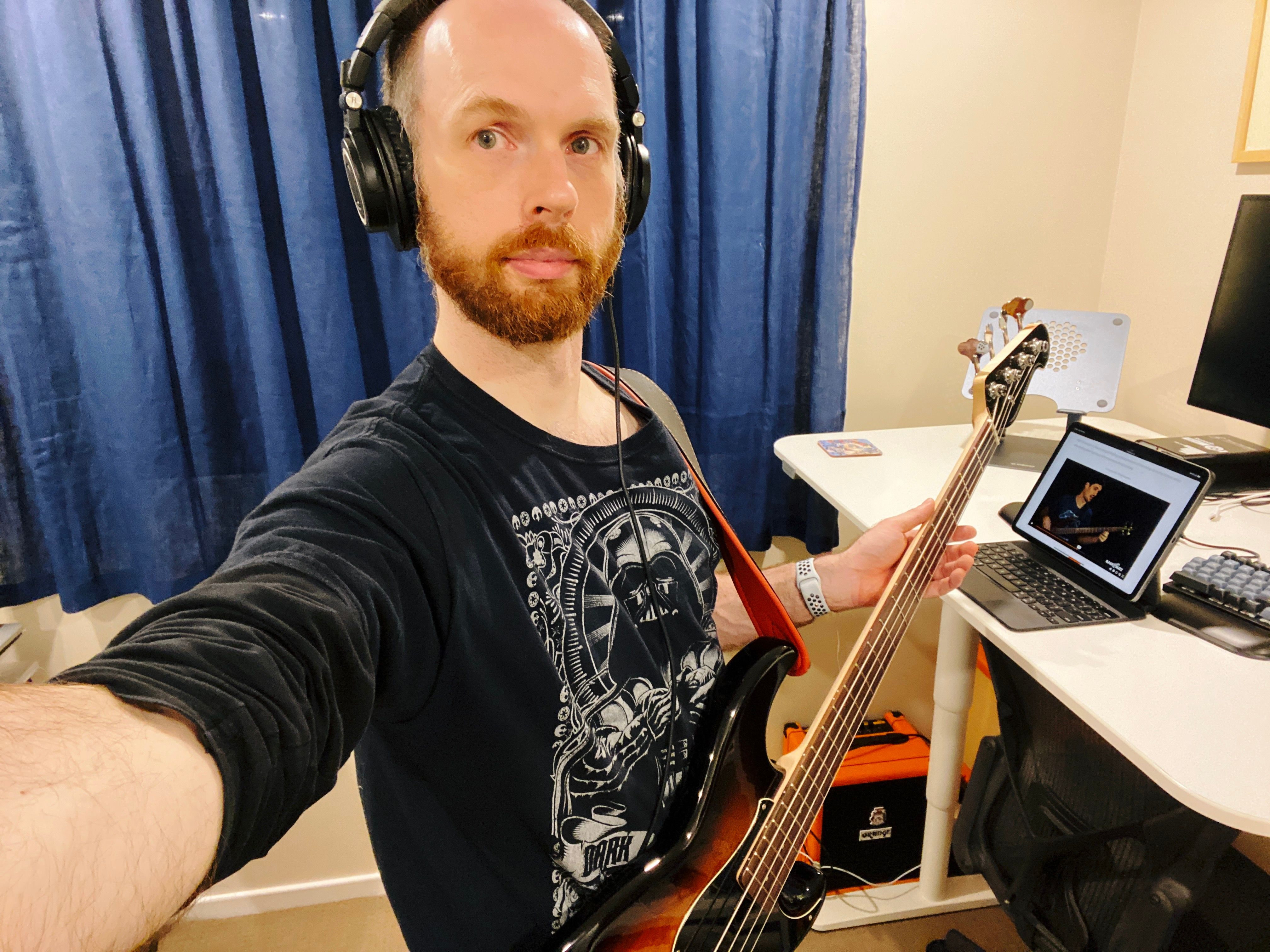 A selfie of me, a white man with short brown hair and an increasingly-scruffy-looking red beard, wearing a black Darth Vader t-shirt. I'm wearing a set of headphones and am holding an extremely handsome-looking bass guitar, a Yamaha BB-434 in Tabacco Sunburst (the colour starts in the middle of the body as a rich deep orange/red colour and fades to black as it gets towards the edge of the body). My iPad is sitting on the standing desk next to me, paused in the middle of a lesson from Josh Fossgreen's "Beginner to Badass" bass guitar course (highly recommended if you're interested in learning bass).