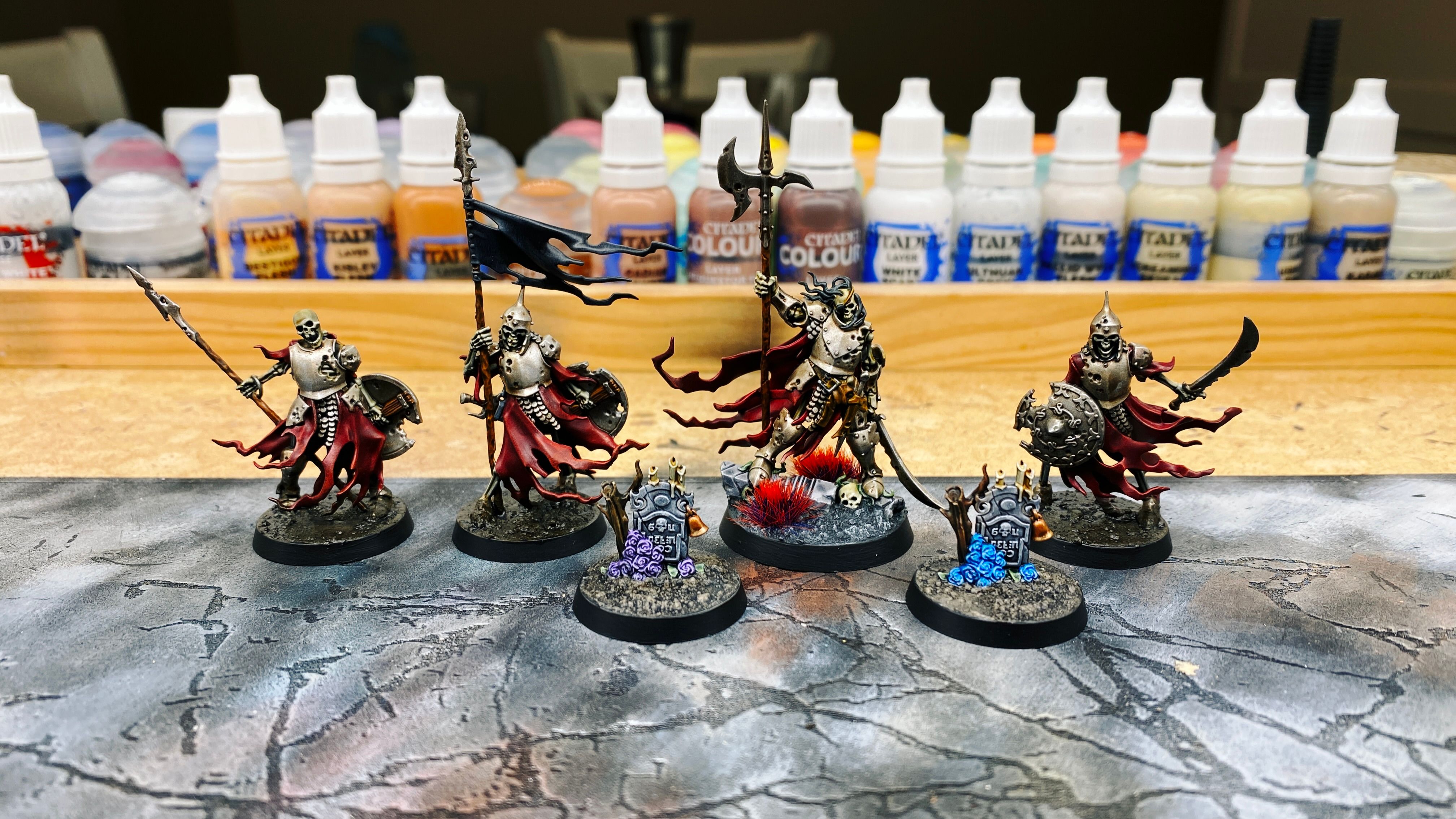 A photo of six painted miniatures, four are skeleton warriors and there's two gravestones sitting on their own bases too. The skeletons all have ripped and torn red cloaks and rusty-looking armour, one of the skeletons is holding a black banner on a long pole, and one is clearly a boss because he's larger with a bigger weapon. The gravestones have a pile of flowers sitting next to them, one blue, one purple.
