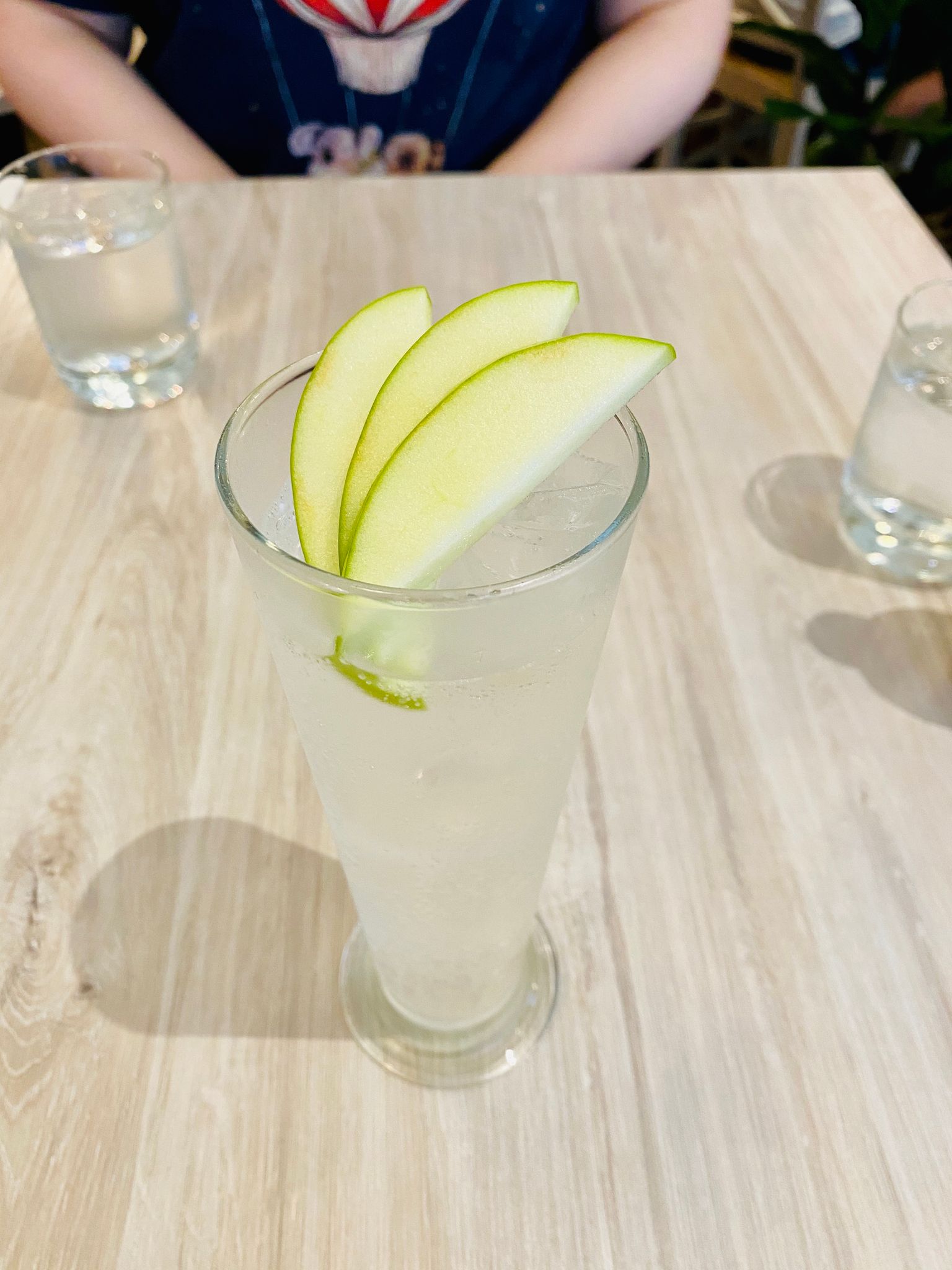 A tall glass of apple cider with ice in it, and three half-slices of apple garnishing the top of it.
