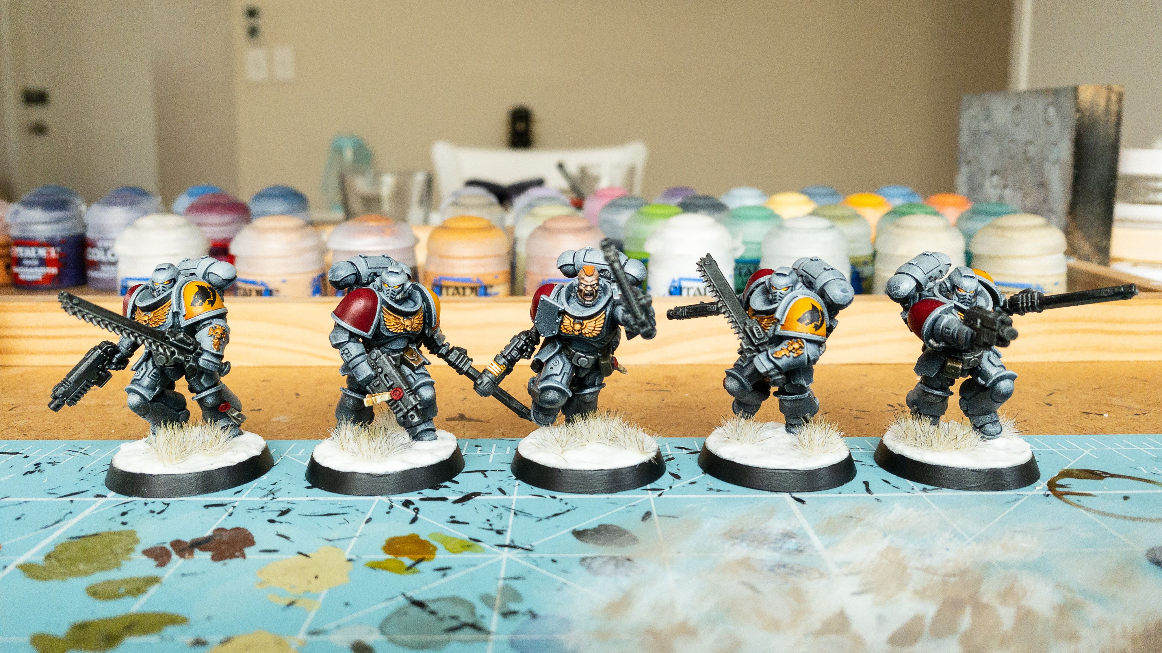A photo of five Space Marines, heavily-armoured warriors all wielding pistols and chainswords. They're painted a blue/grey colour, one shoulder pad is a rich yellow and the other is a dark red. The sergeant in the middle has no helmet on and I was able to paint his pupils, and for the other four with helmets on, their eye slits go from red on the outside to yellow on the inside. They're on snowy bases and the bases have little wintery grass tufts on them.
