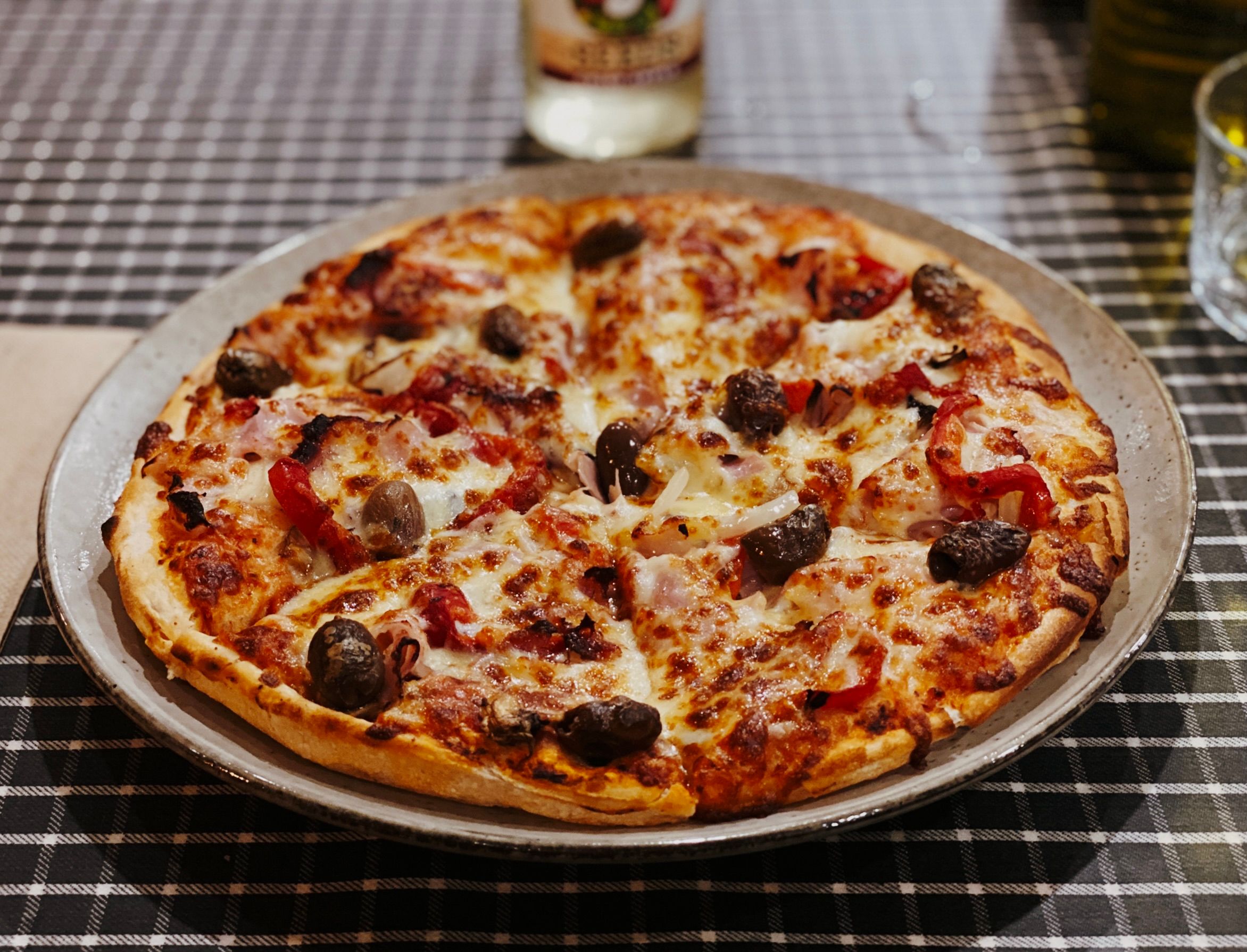 A photo of a pizza sitting on a plate. Just visible in places under the cheese are pieces of ham and onion, and on the top it has sun-dried tomatoes and olives.