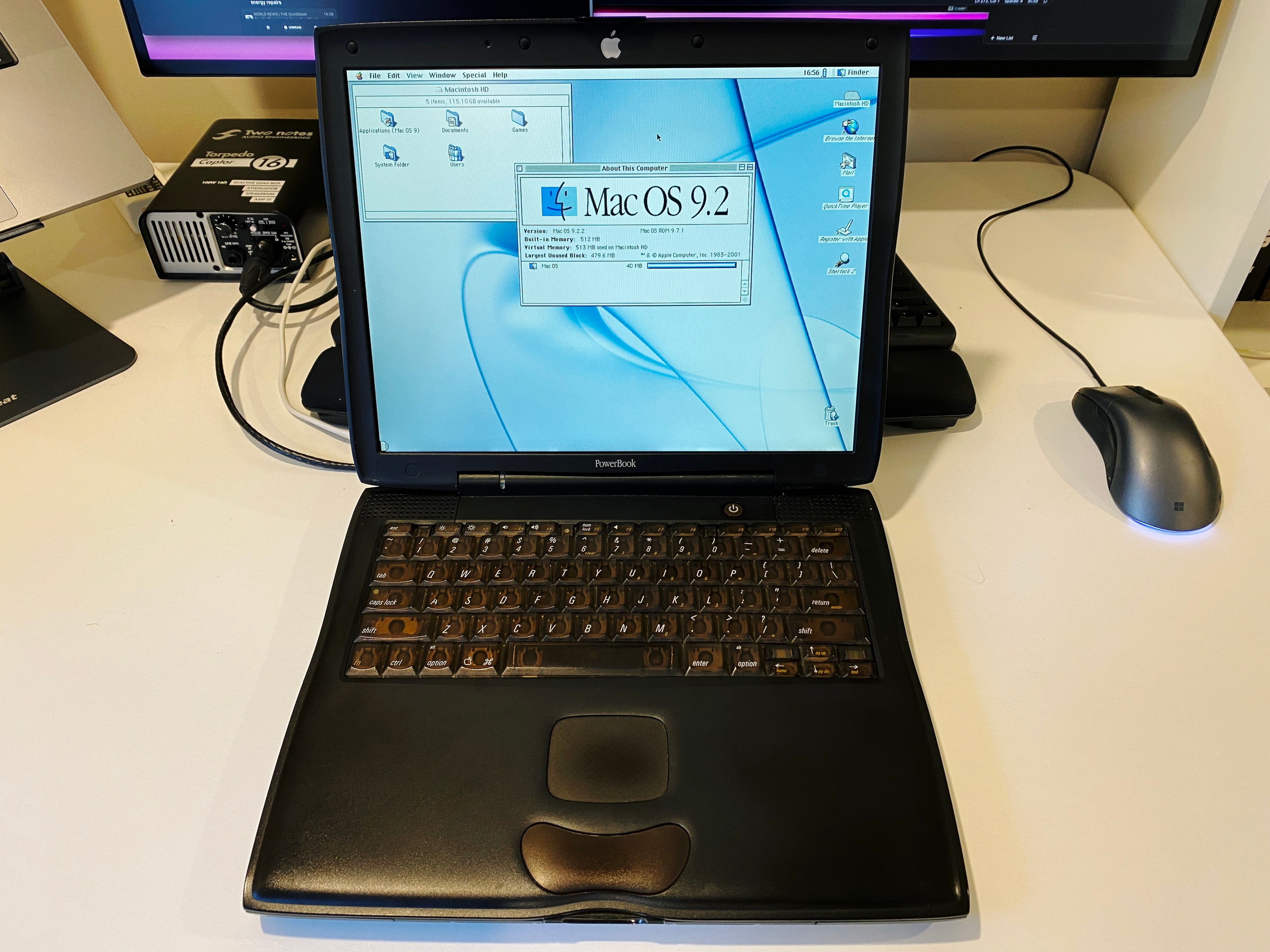 A photo of a black PowerBook G3 sitting on a desk, booted to the Mac OS 9 desktop. The machine is big and chunky, but also has subtle curves to it, and the trackpad is HILARIOUSLY tiny compared to modern Macs.