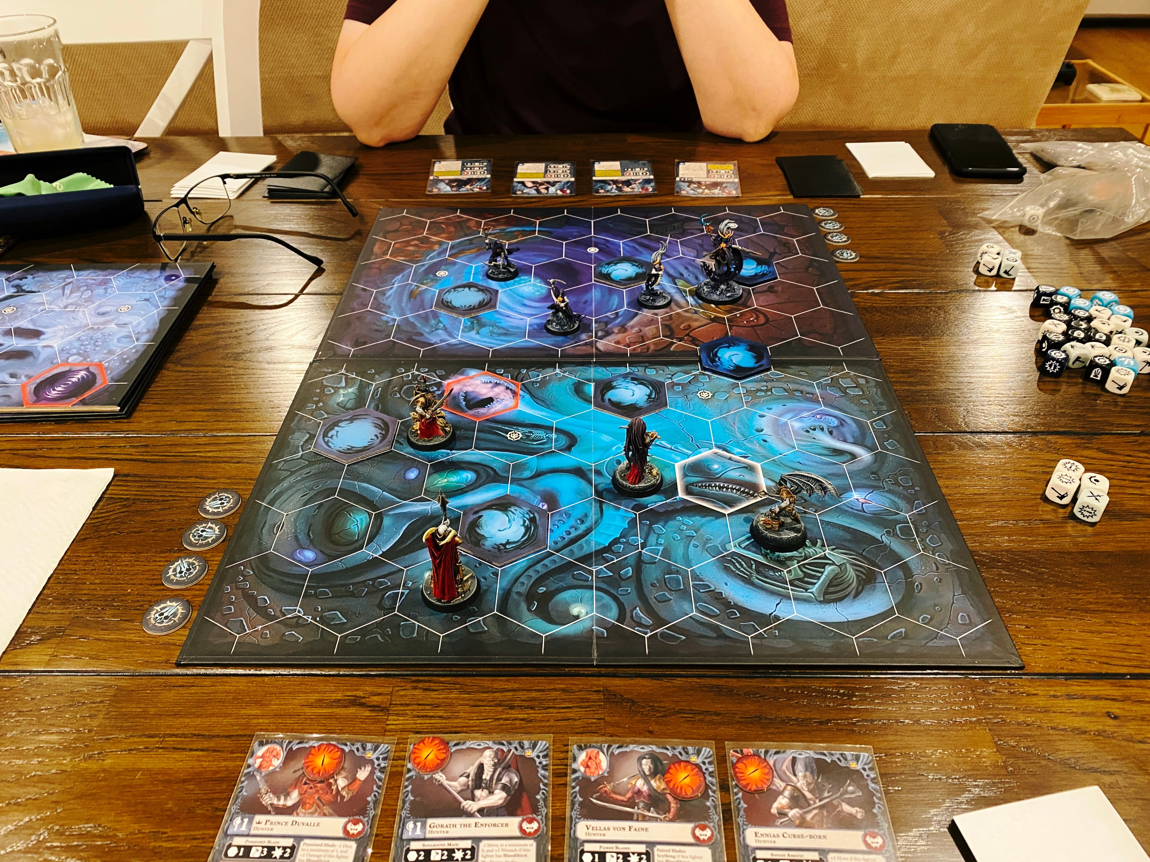 A photo of a two-player game of Warhammer Underworlds. It's a combination miniatures/deck building game played on a board with hexes on it, the artwork on the board looks like it's in a sea cave or something. Closest to the camera are four vampires with red cloaks on, and facing them are four lithe elves painted mostly in deep purples.