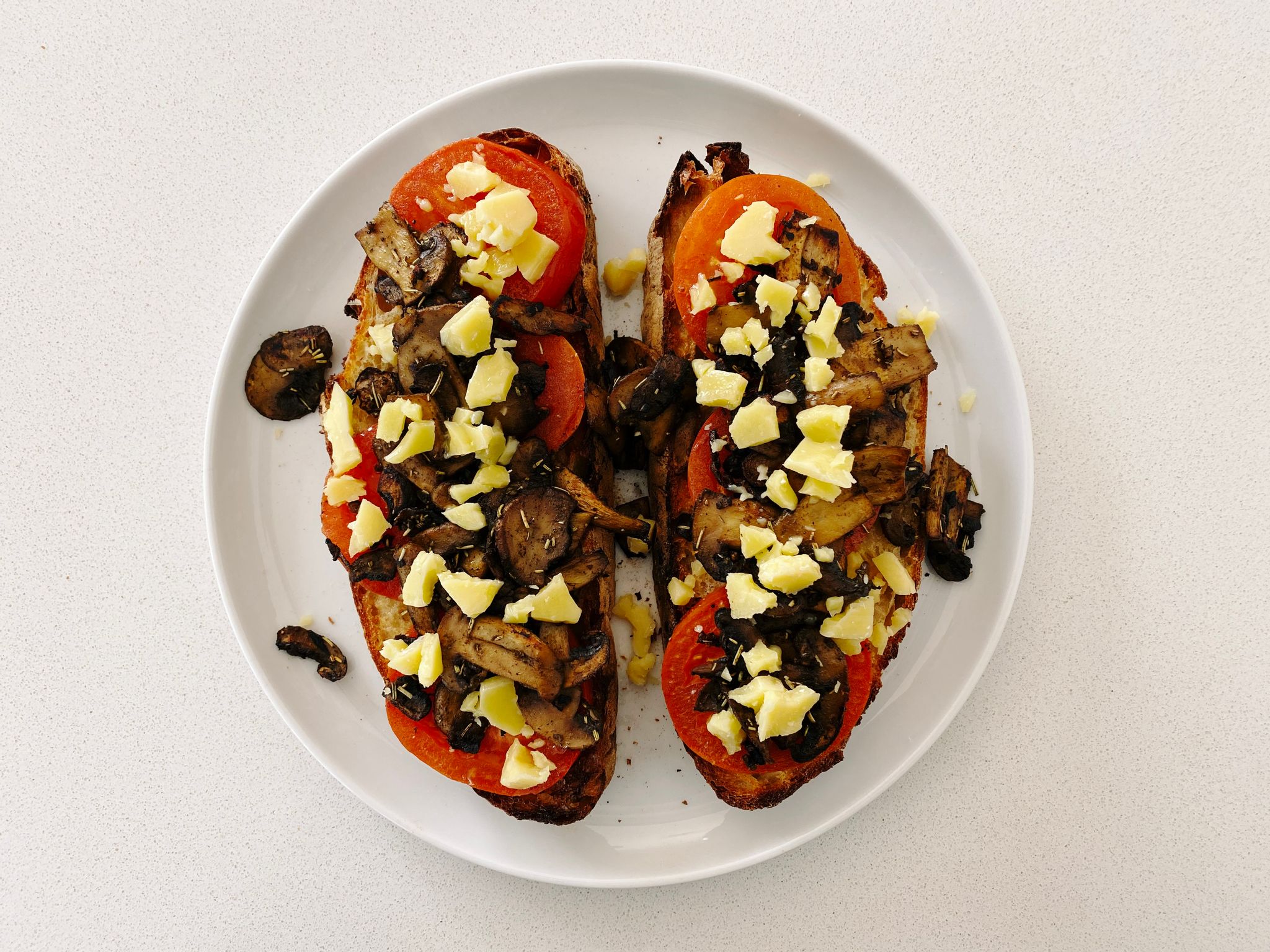 A photo of two slices of homemade bread sitting on a plate, topped with thick slices of tomato with mushrooms on them, and small pieces of crumbled up cheddar cheese atop it all.