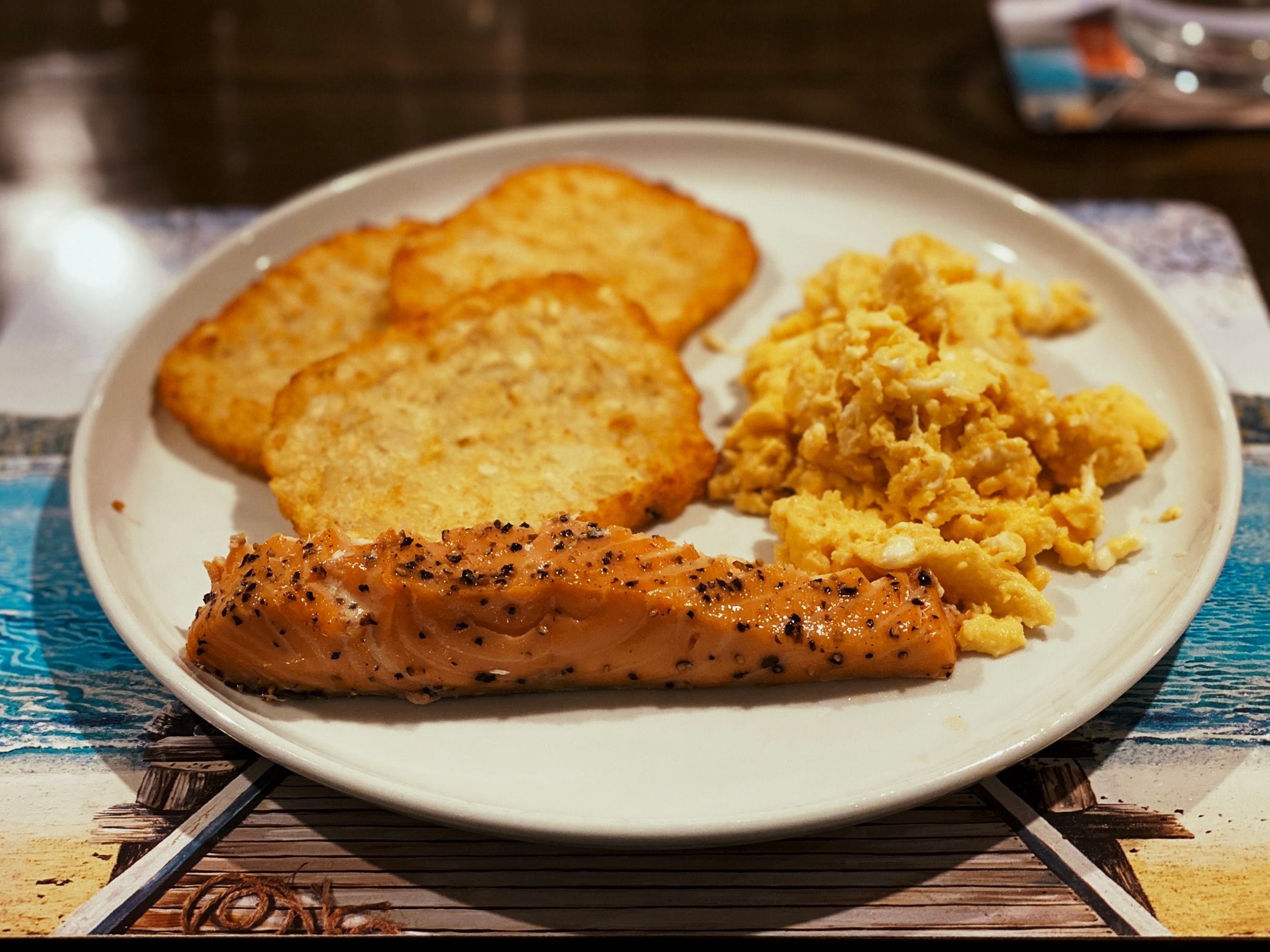 A slightly arty photo (i.e. taken with portrait mode so there's a nice narrow depth of field) of a slice of smoked salmon sitting on a plate alongside three golden-brown hash browns and a pile of scrambled eggs.