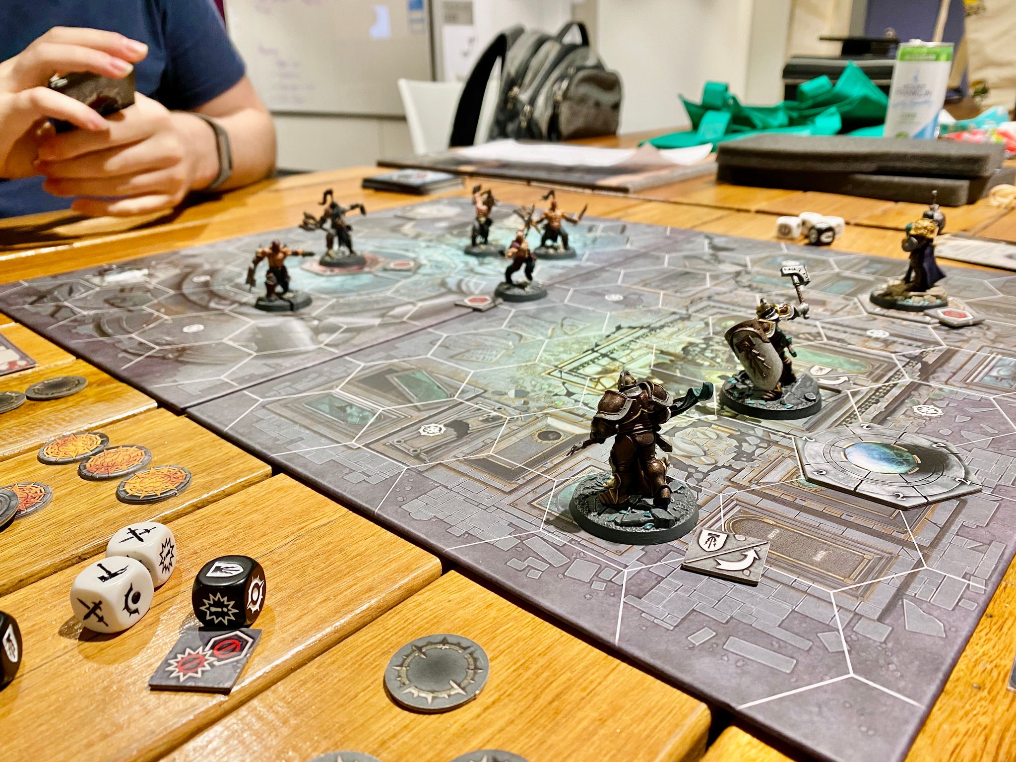 A photo taken from table level, looking across the game board from Warhammer Underworlds: Shadespire. At the right are three heavily-armoured warrior miniatures in full plate, facing them at the left are five human berserker-types wearing basically no armour at all. The table is covered in tokens and dice.