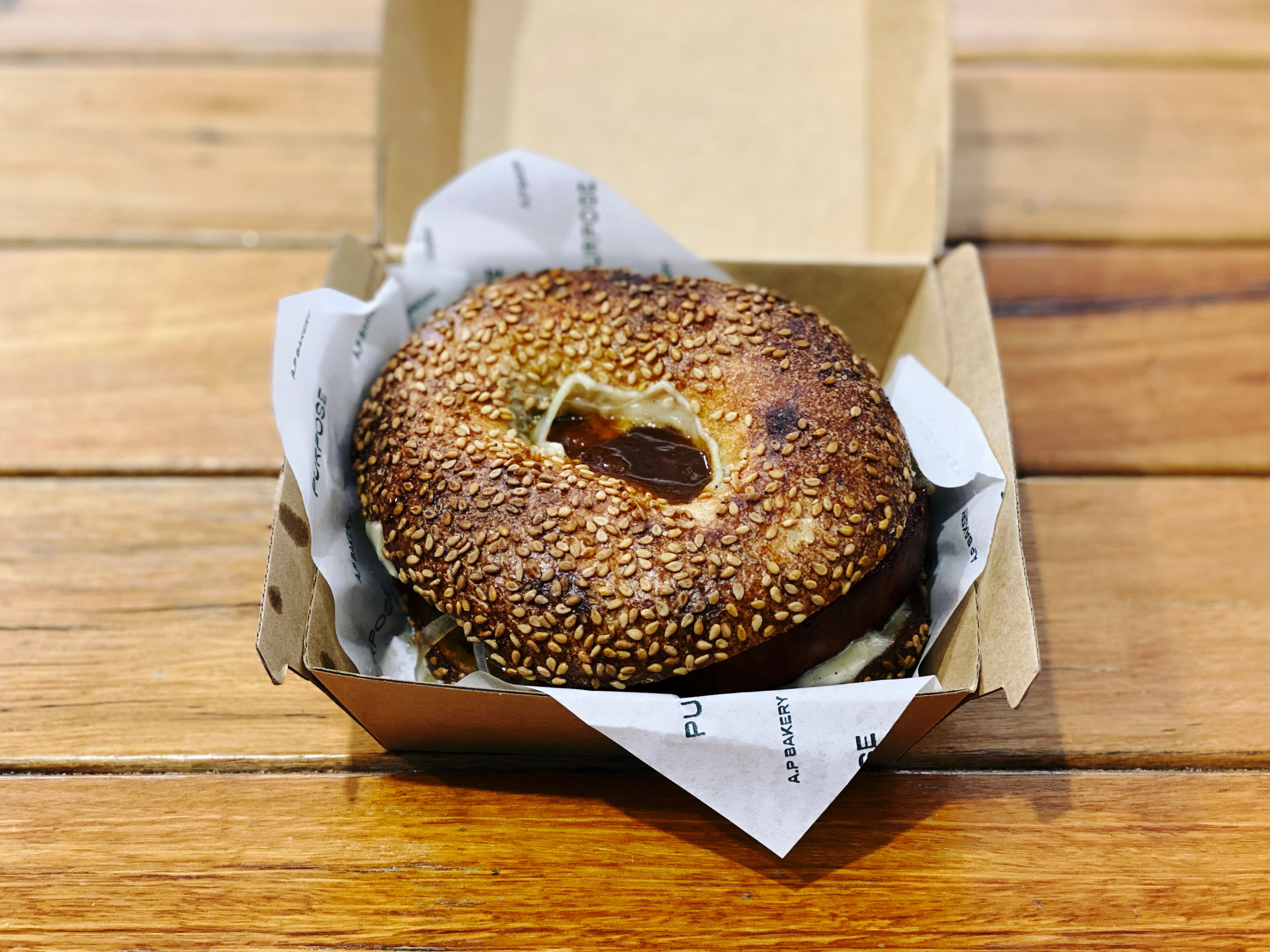 A photo of a sesame bagel sitting in a cardboard takeaway container with some cheese and sauce visible through the hole. It's hard to see in the photo but the piece of chorizo was nearly as large as the bagel and probably would have been close to a centimetre thick.