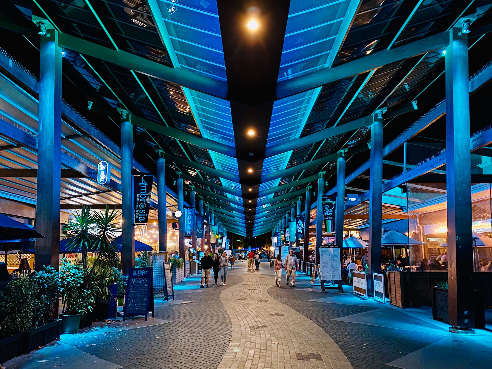 A night time photo looking down an pedestrian "street" with restaurants on either side and bright turquoise lighting along the ceiling/roof area.