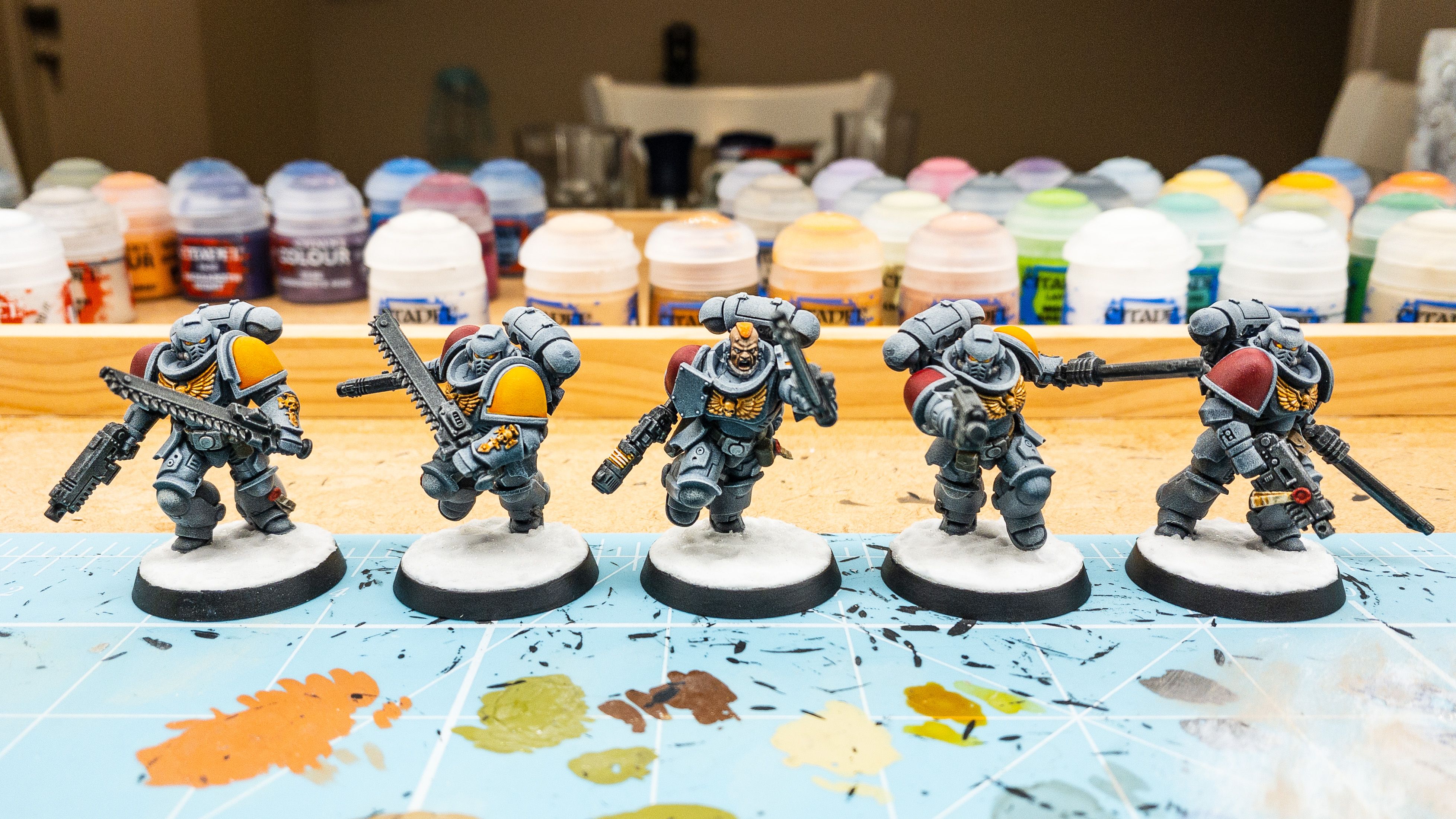 A photo of five Space Marines, all wielding pistols and chainswords. They're painted a blue/grey colour, one shoulder pad is a rich yellow and the other is a dark red. The sergeant in the middle has no helmet on and I was able to paint his pupils, and for the other four with helmets on, their eye slits go from red on the outside to yellow on the inside.