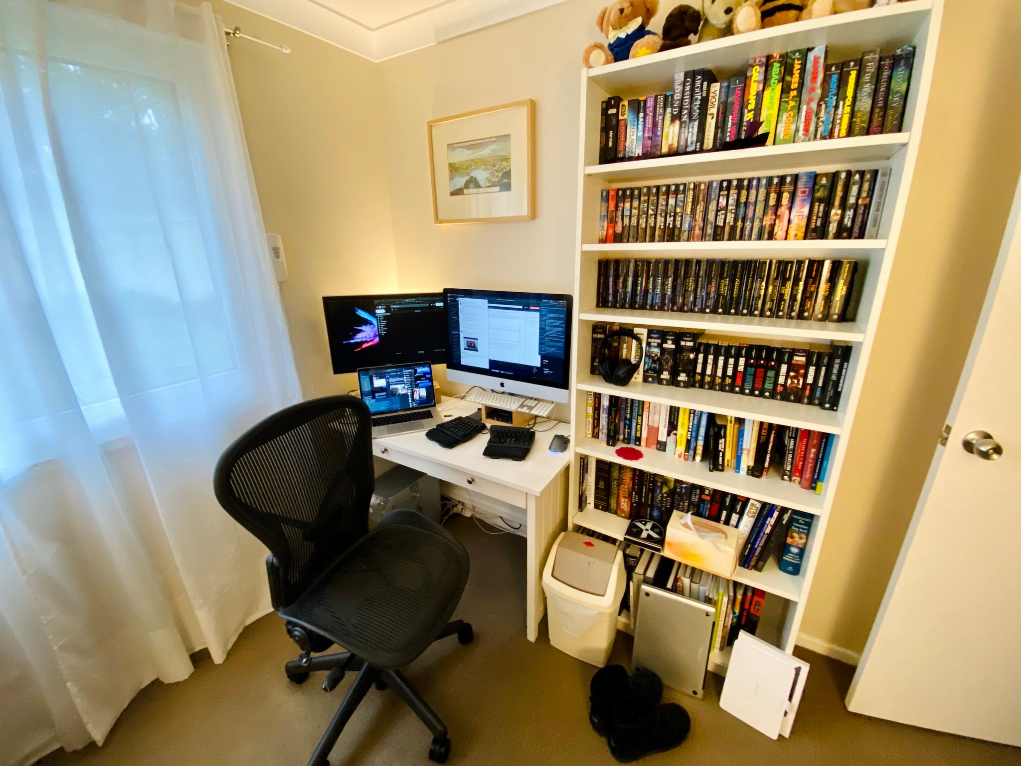 A photo of a computer desk in the corner of a room next to a bookshelf full of books. The desk has an iMac, a 24" monitor, and a MacBook Pro sitting on it.