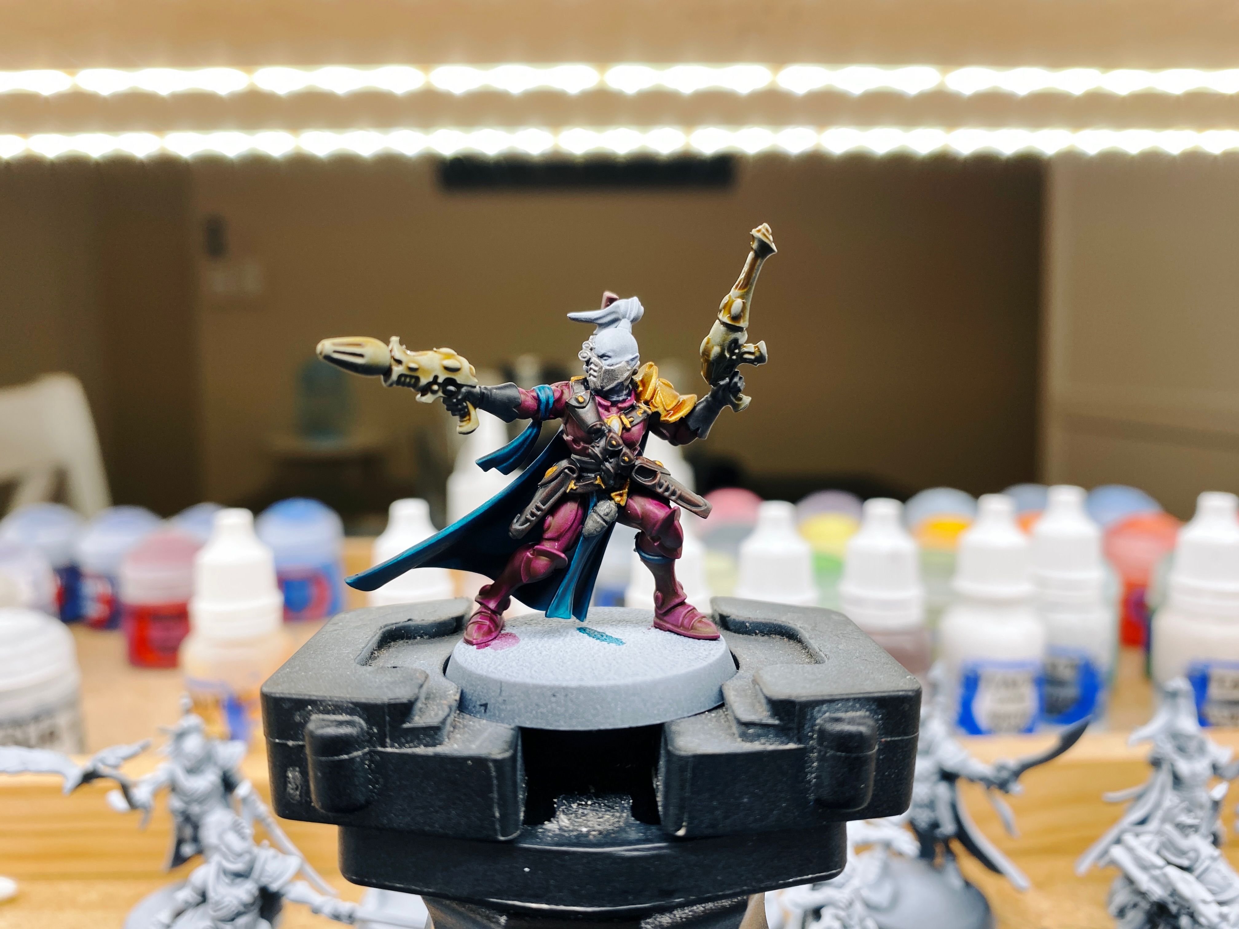 A photo of a partially painted Aeldari miniature (think "space elf"). She's wearing elegant fuchsia-coloured armour with a dark turquoise cape flowing behind her and a bunch of dark leather pouches across her chest, and is holding two bone-coloured alien-looking pistols, one pointing like she's about to shoot and the other one behind her pointing upwards. She's got a cool-looking rebreather on that has an integrated targeting set up over one eye, and she's got a bald head except for a big tall top knot.