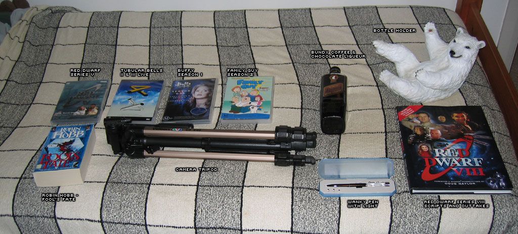 A photo of a selection of DVDs, a couple of books, a bottle of alcohol, an alcohol bottle holder that is the Bundaberg Rum bear lying backwards so it can hold the bottle with its arms and legs, and a camera tripod, all sitting on a bed.
