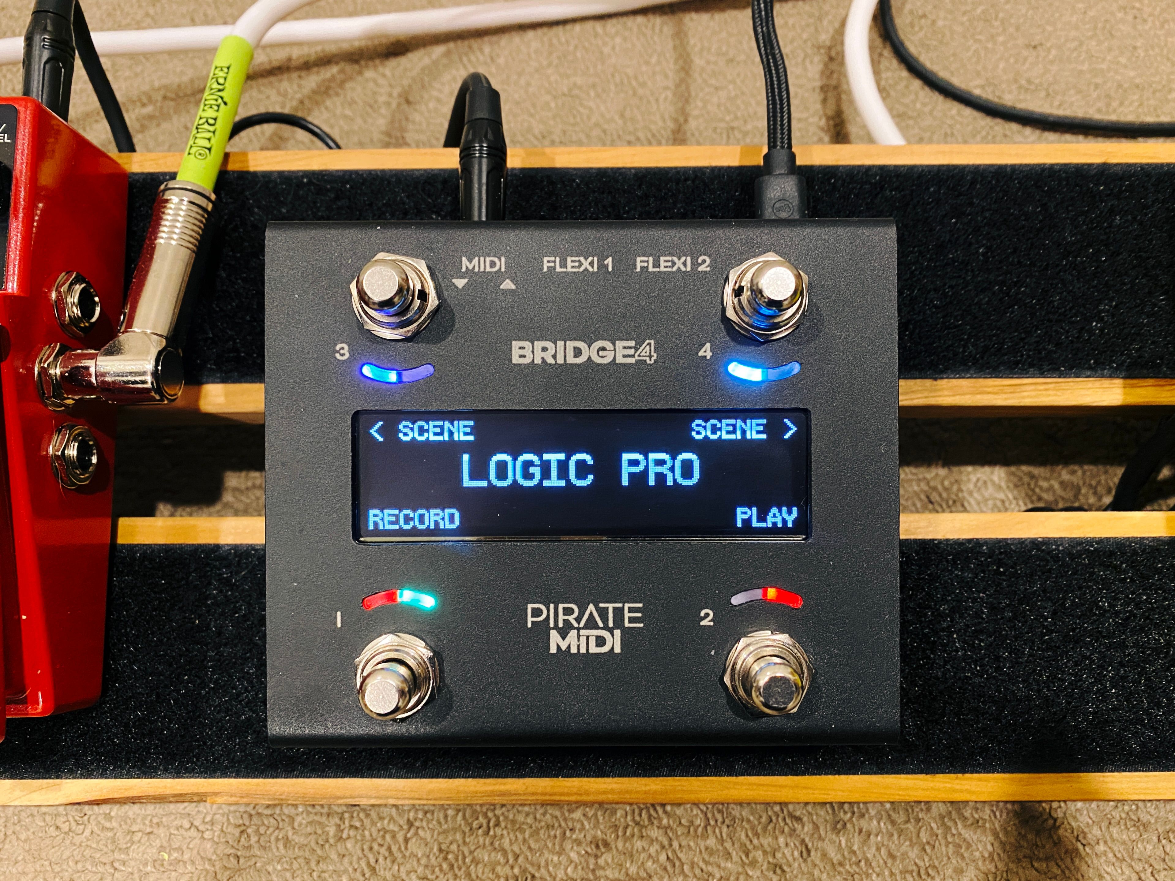 A photo of a black metal box with four silver push switches and an LCD display in the middle. The display says "LOGIC PRO" in the middle with "Record", "Play", "Previous Scene" and "Next Scene" in the corners corresponding to each switch.