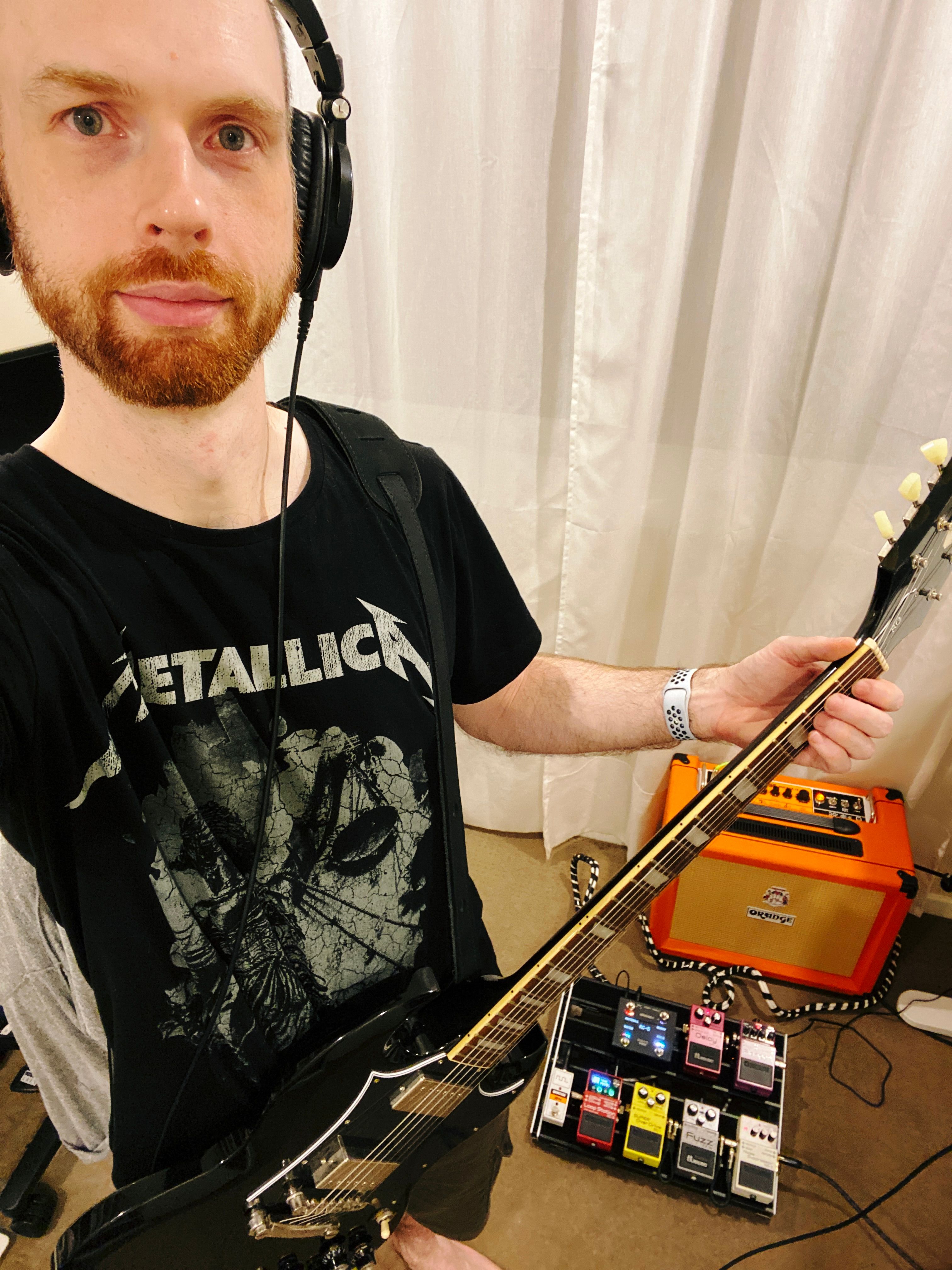 A selfie of me, a white man with short hair and a short red beard (and a very receding hairline) wearing a black Metallica t-shirt and holding a black electric guitar. In the background on the floor are my guitar amp and pedal board.