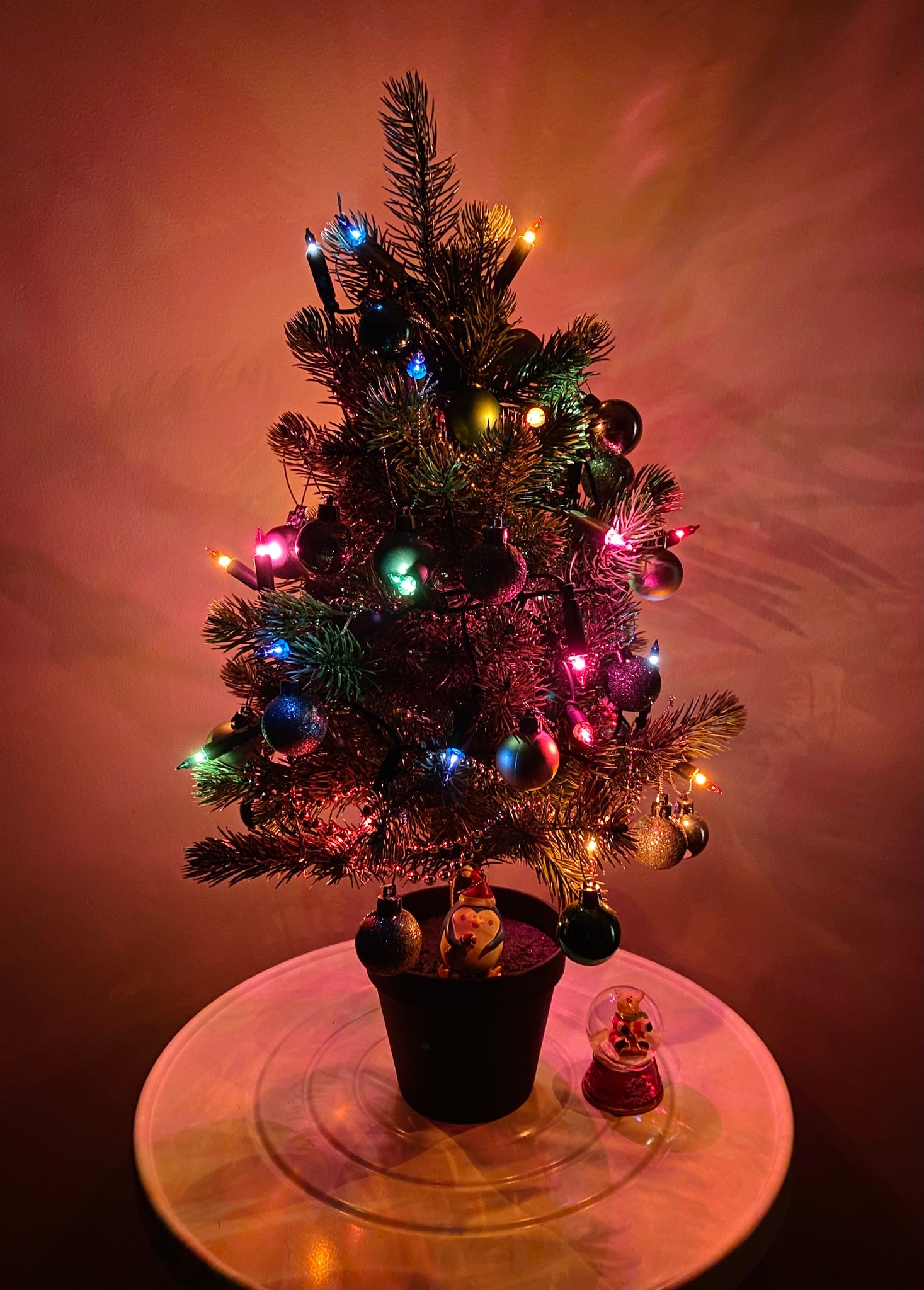 A photo of a small (~40cm high) plastic Christmas tree in its own fake pot, covered with tinsel and lights, with a penguin in Santa hay at the base and a Santa snow globe next to it.