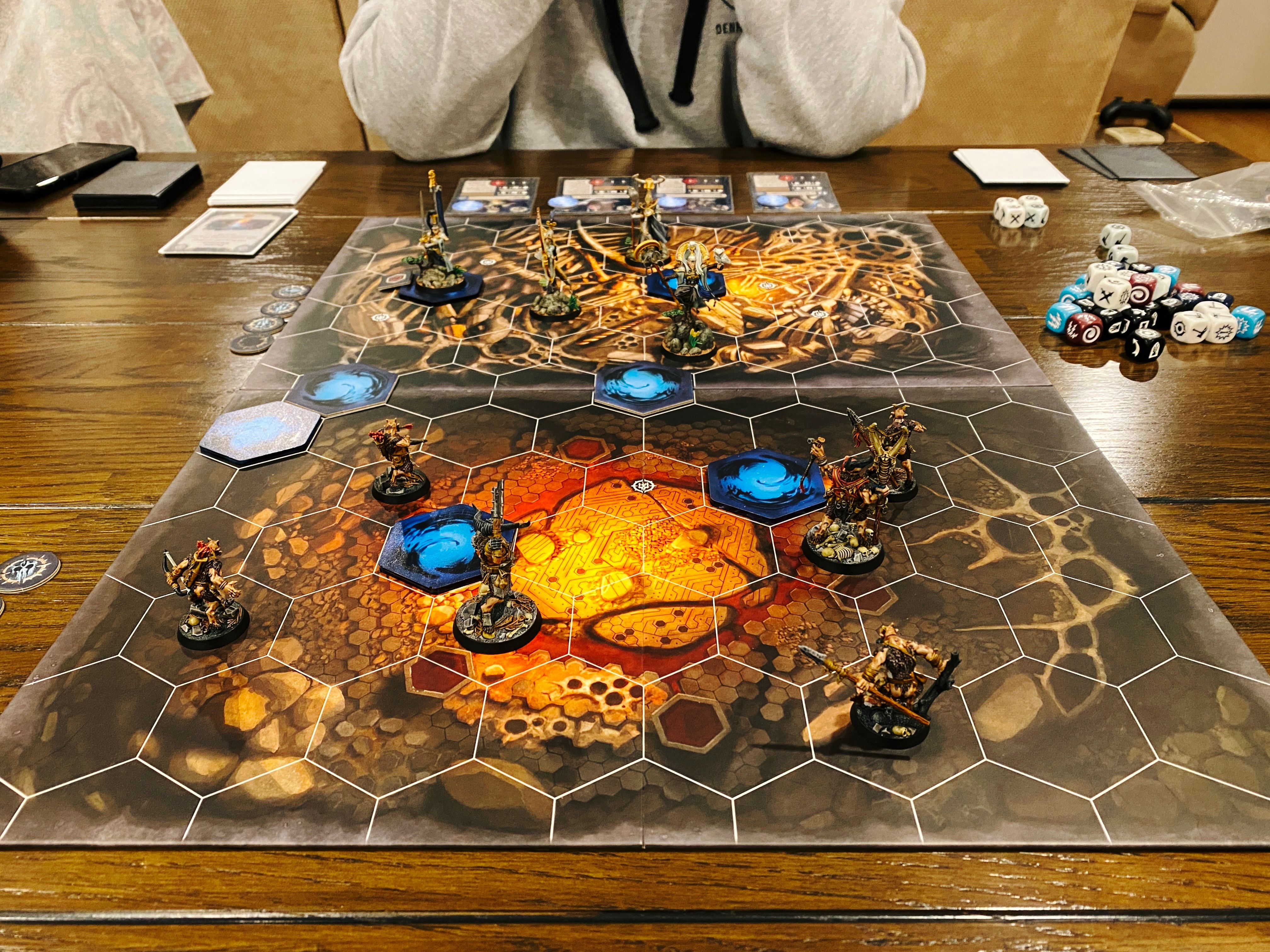 A photo of Warhammer Underworlds game board. The board itself is covered in hexes with the artwork underneath looking like deep within a cave, and at the front closest to the camera are six savage beastmen all yelling and wielding weapons like they're about to charge, and facing them across the board are four lithe and elegant aelves.