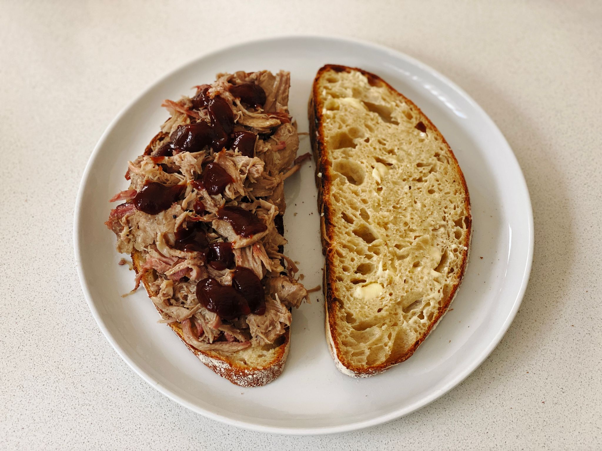 A photo of two slices of the aforementioned bread sitting on a plate, one of the pieces has a ton of pulled pork on it and dollops of barbecue sauce on top.