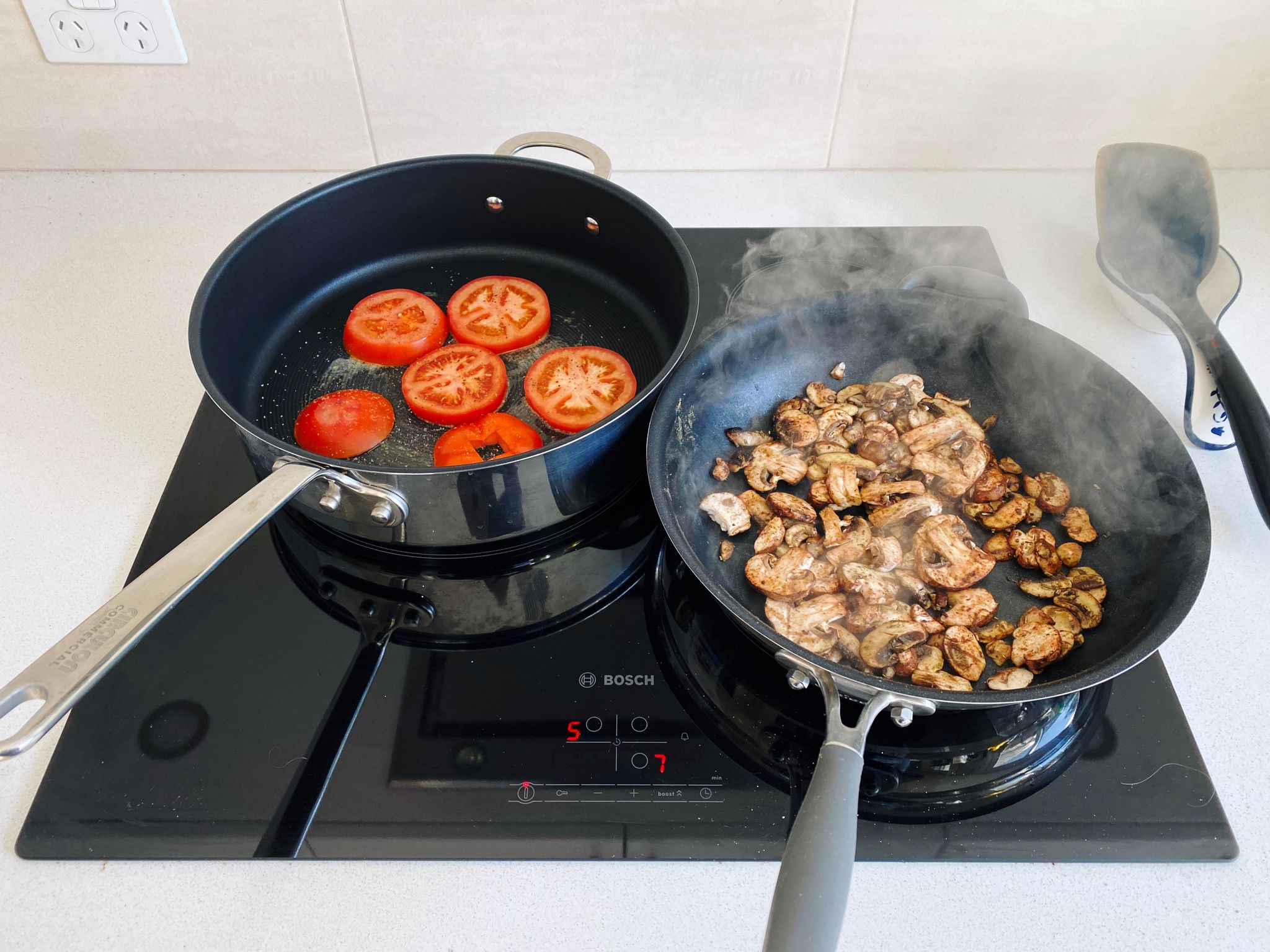 A photo of two frying pans on a stove, one with sliced tomatoes in it and the other with mushrooms.