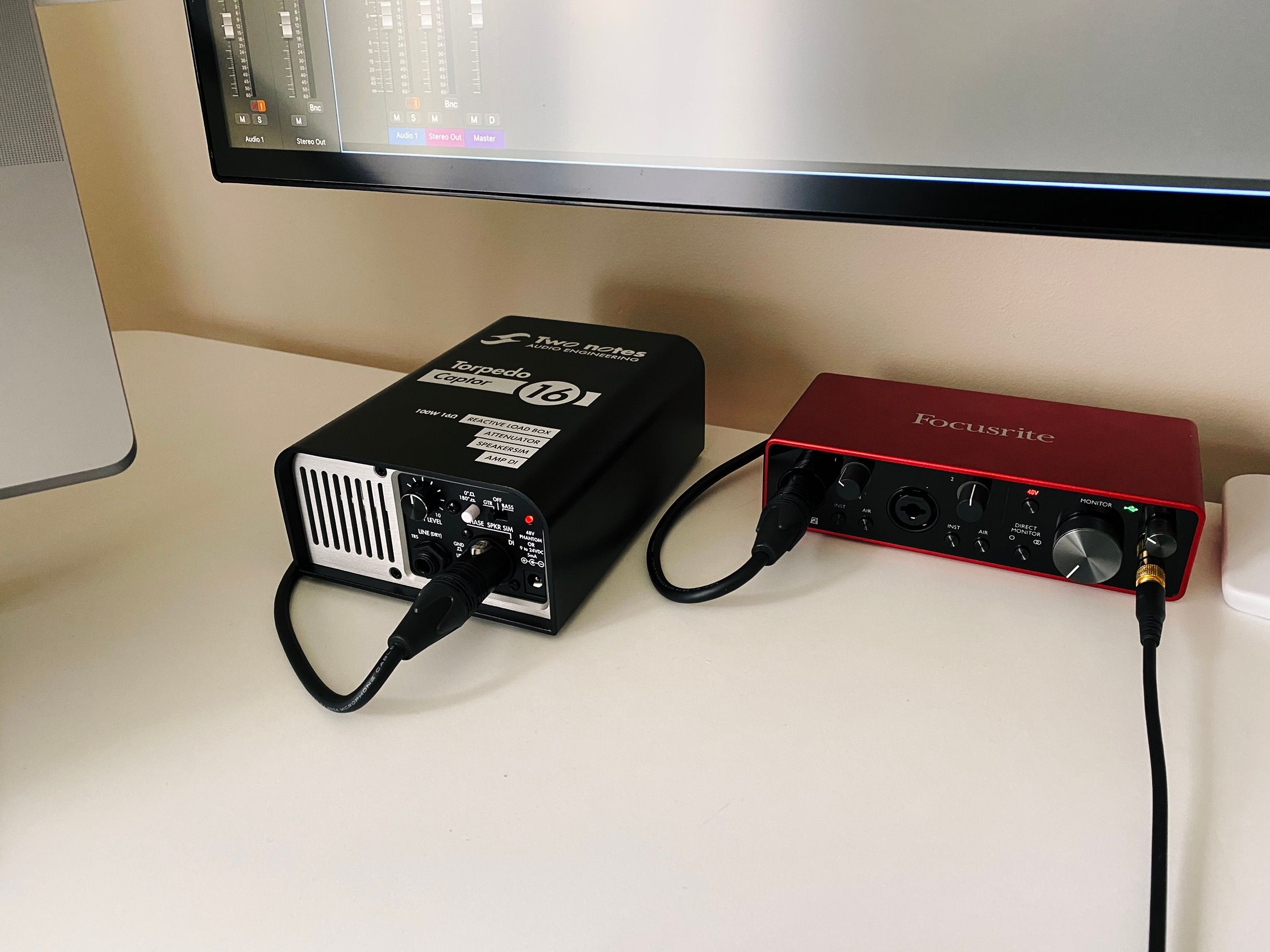 A photo of a Two Notes brand "Torpedo Captor" guitar load box. It's a big chunky black box with dials and switches on the front-right, and a fan intake grill on the front-left. There's a CHONKY-looking XLR cable coming out of it and going into the red Focusrite Scarlett 2i2 USB audio interface next to it, the top and sides of which are a rich deep red metallic colour, and on the front is shiny black with all sorts of dials and sockets on it.