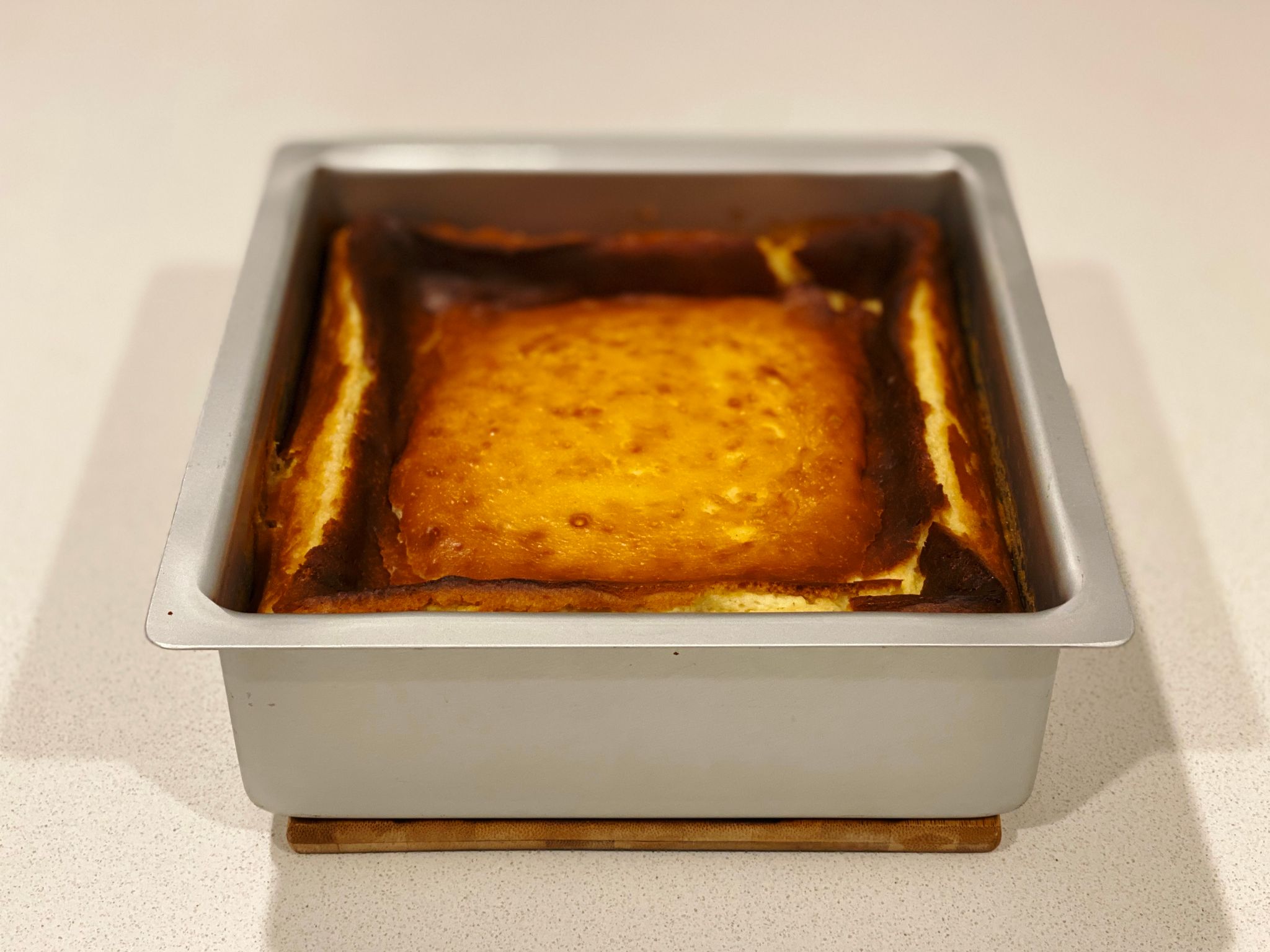 A photo of a tall square baking pan with quite brown-topped cheesecake in it. The edges are higher than the middle, and it looks slightly collapsed. Smells amazing though.