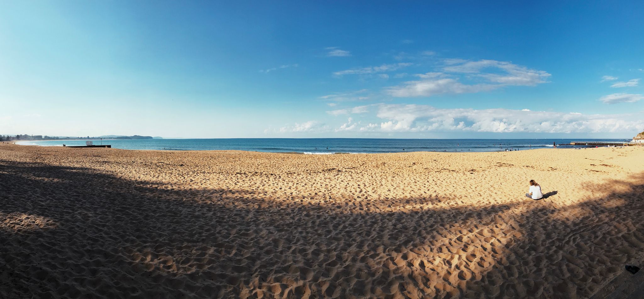A panorama photo of a yellow sandy beach, blue ocean past it, and some clouds towards the horizon in an otherwise blue sky.