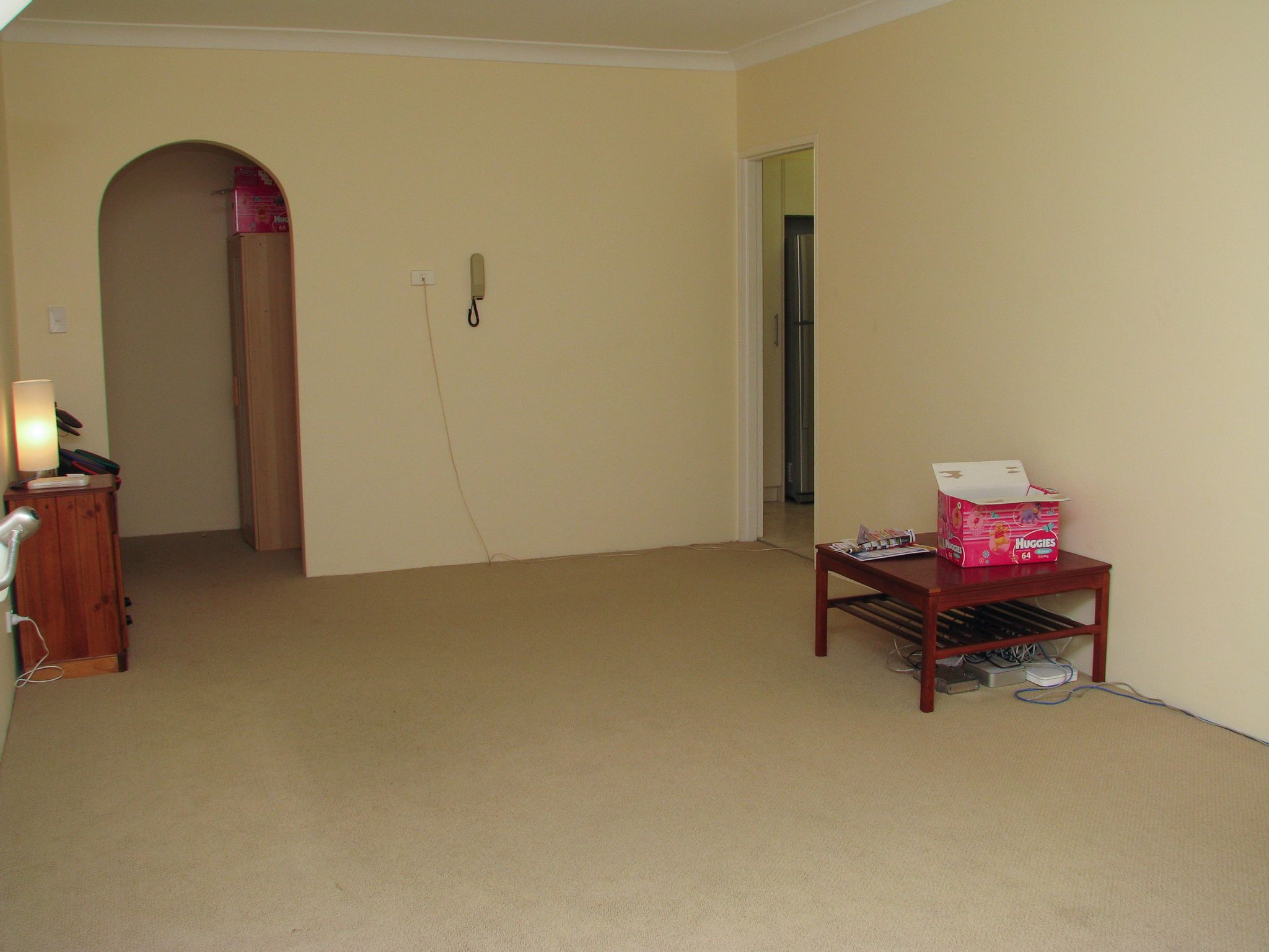 A photo of a lounge room that is totally devoid of furniture except for a coffee table against the wall with some networking gear underneath it and a box of "Huggies" nappies on top, and a bedside table.