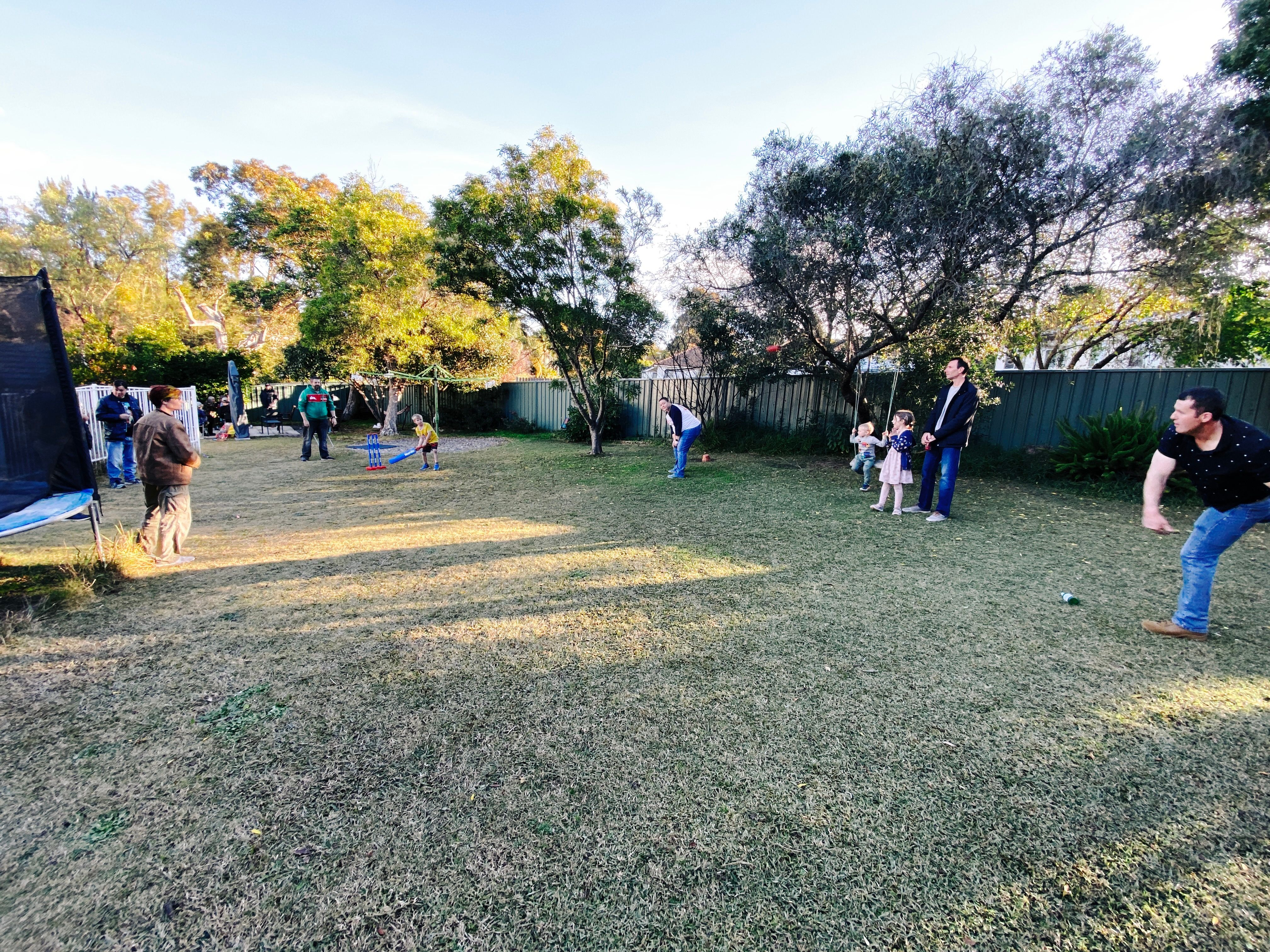 A photo of a grassy backyard with a set of plastic cricket stumps set up, a 6 year-old holding a plastic cricket bat, and a man mid-bowl just in frame at the right. There's a number of other adults and children standing around fielding and just generally watching.