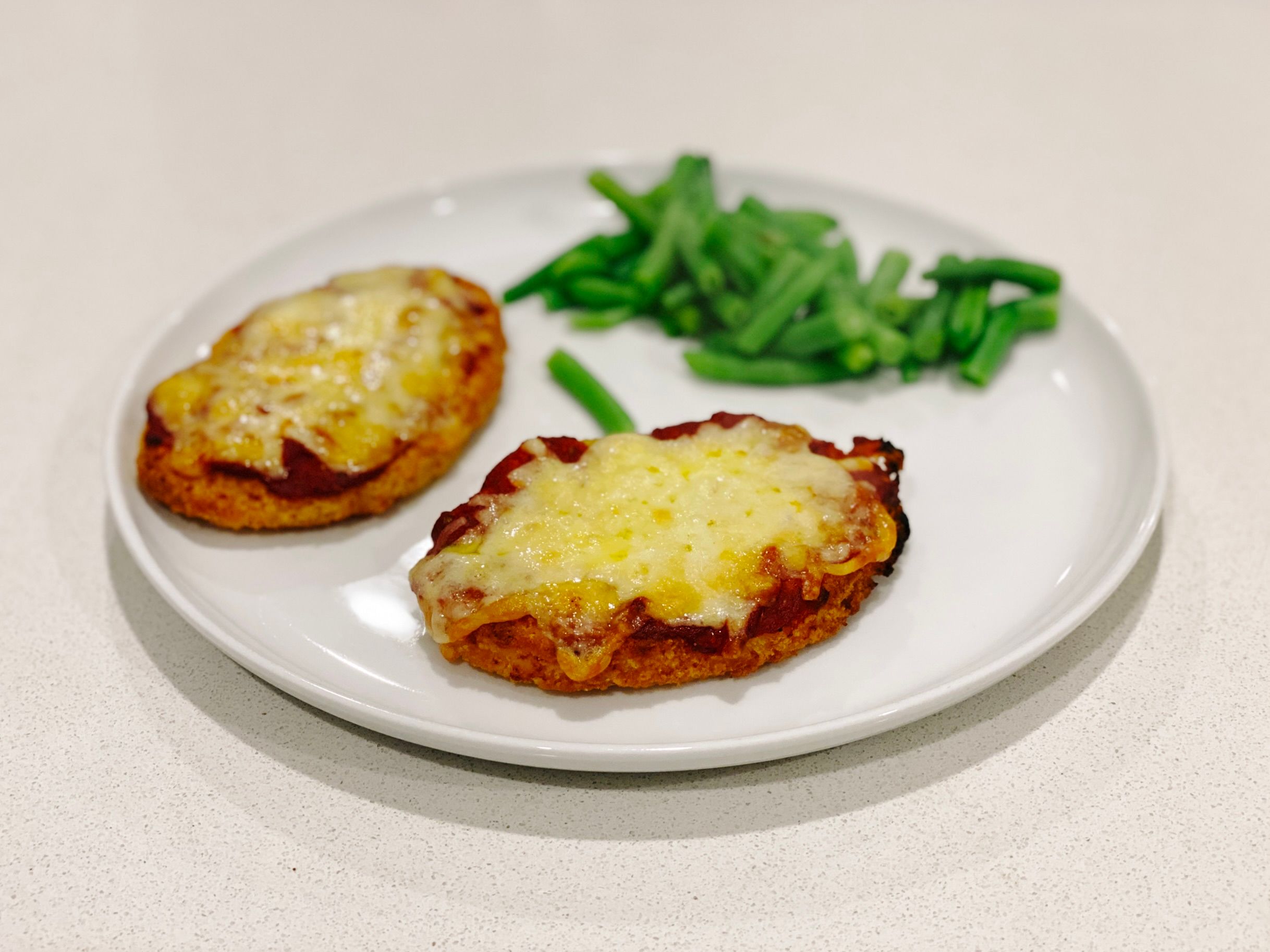 A photo of two pieces of chicken schnitzel with tomato paste and cheese on top, sitting on a white plate with some green beans next to them.