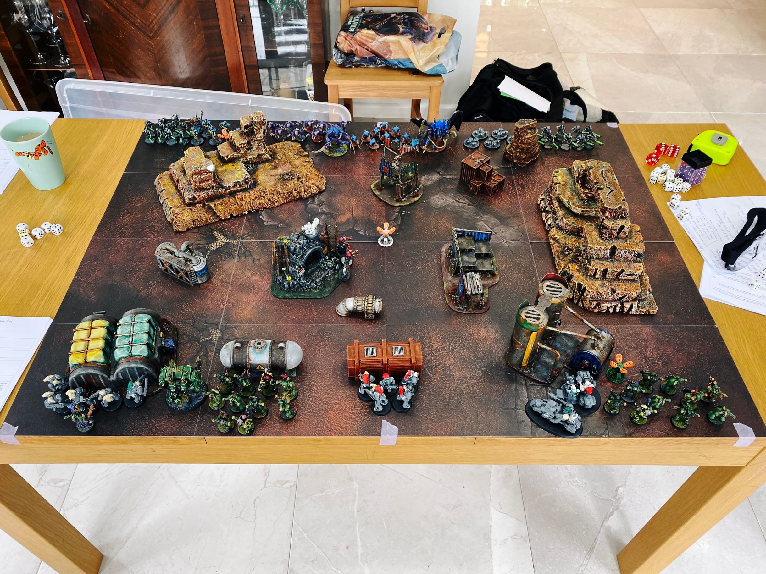 A photo of a table with lots of home-made terrain on it, all either rocky or scrap-looking metal constructs. On this side are Chaos Space Marines, painted in green, next to regular Space Marines which are shamefully unpainted (and also held together with Blu-tac). On the other are Tyranids and Necrons.