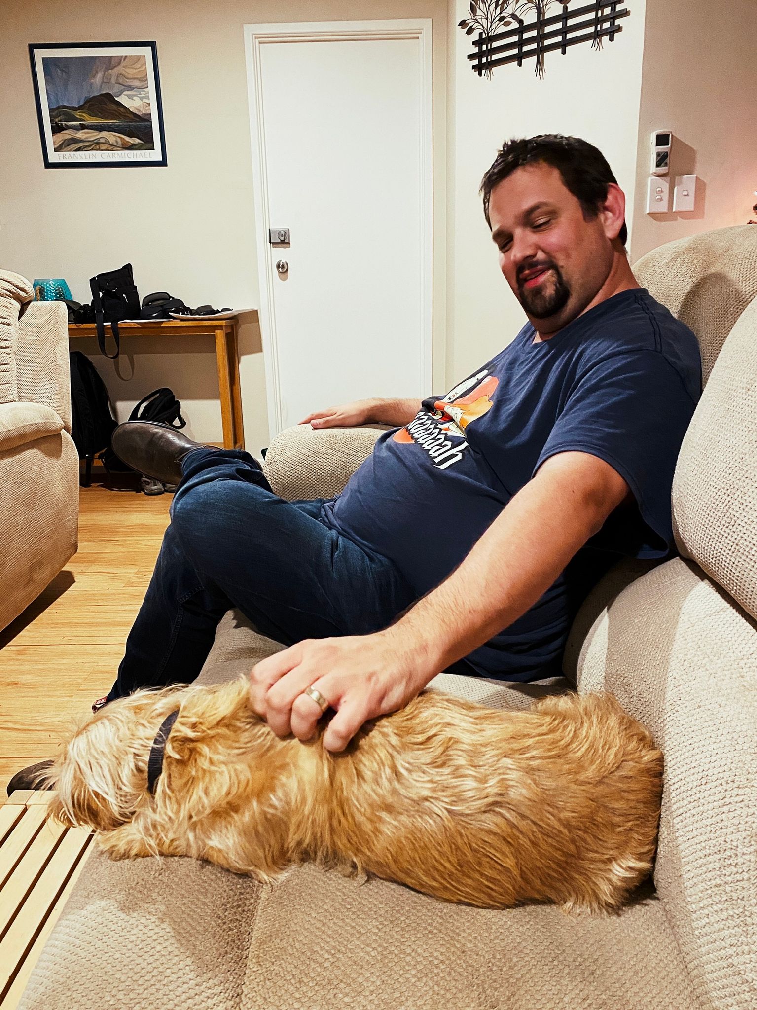 A photo of a white man in a t-shirt sitting on a lounge, giving pats to a small scruffy blonde dog that's lying down next to him.