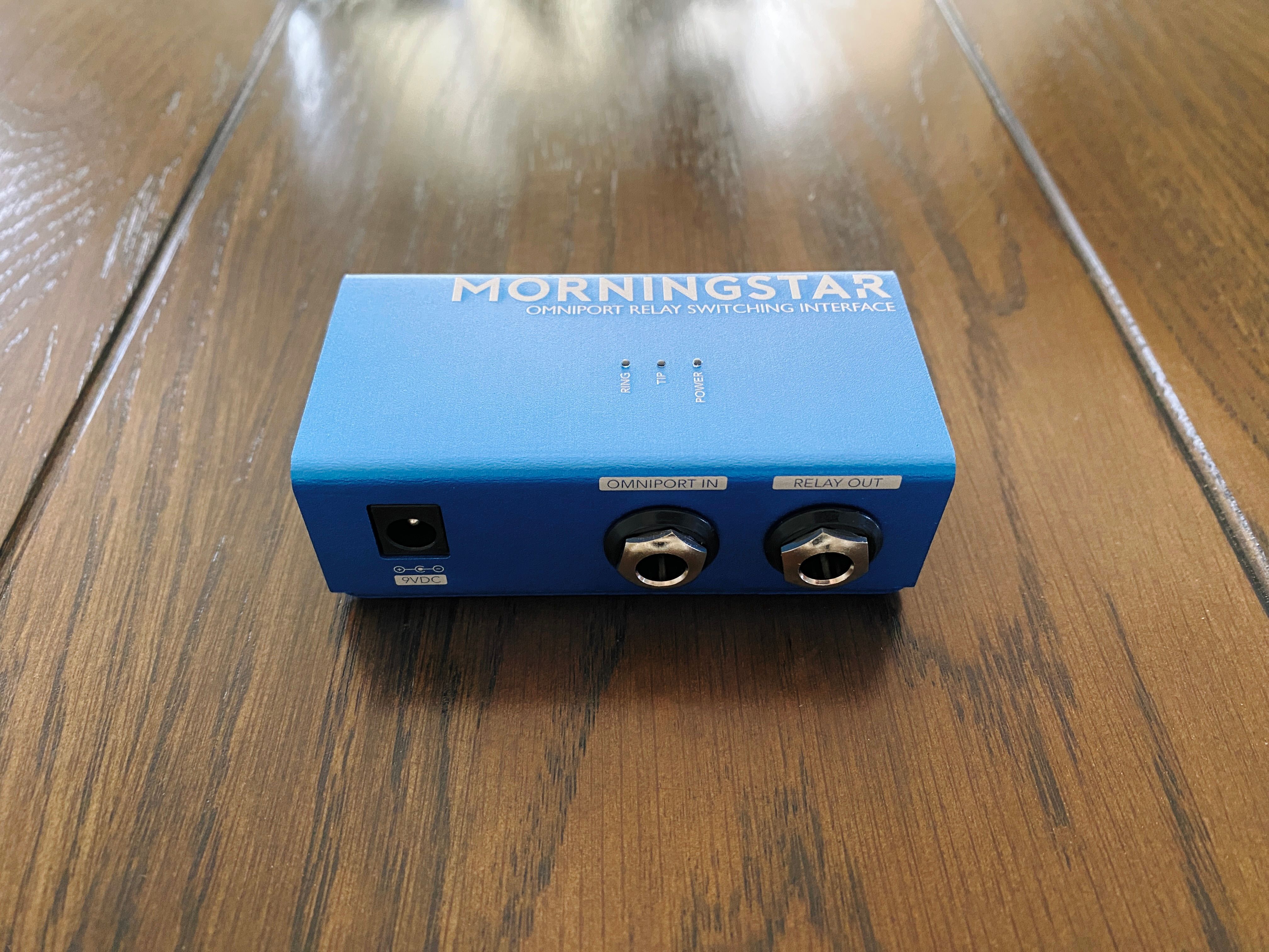 A photo of my Morningstar relay switching interface. It's a small blue metallic box with two TRS jacks and a DC in jack on the front, and three LEDs on the top marked "RING", "TIP", and "POWER".