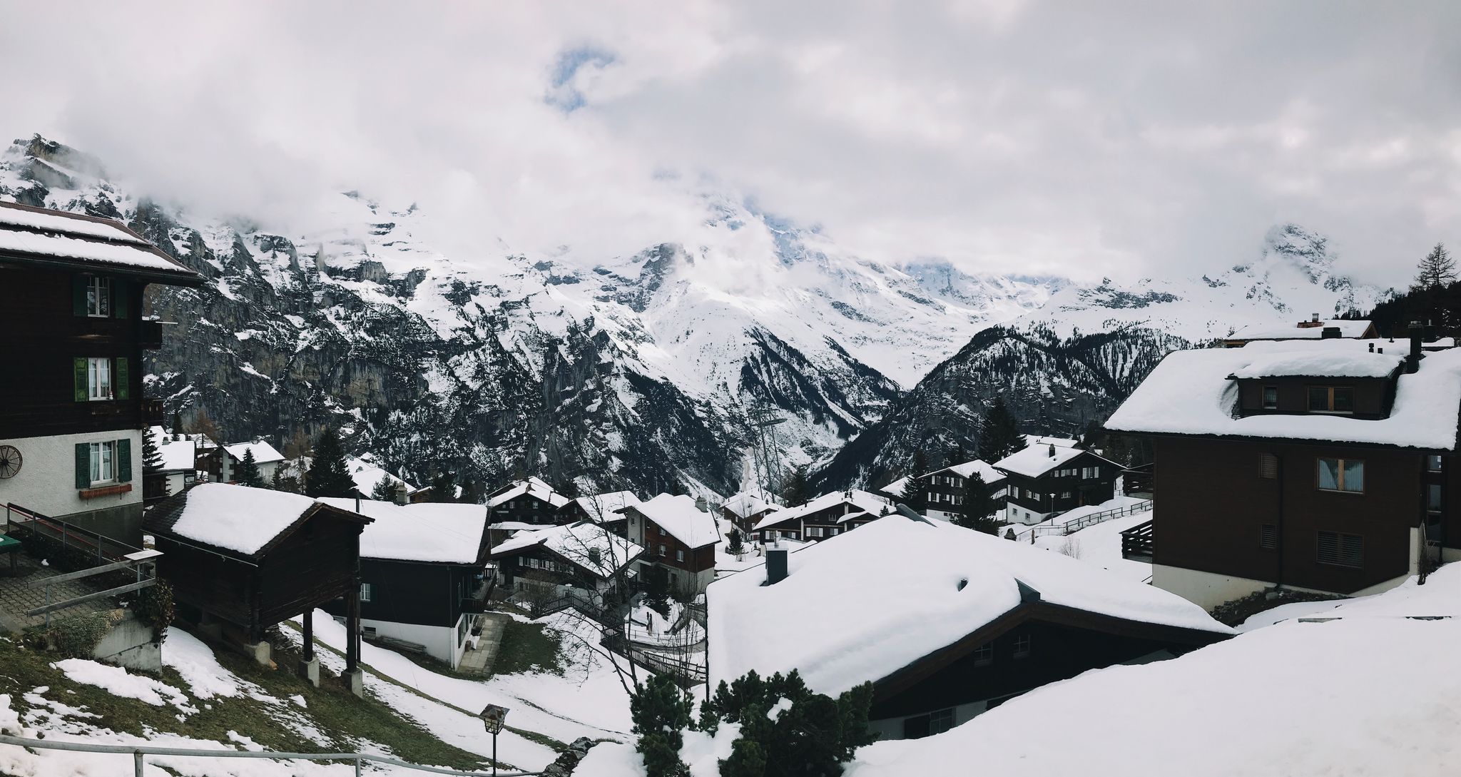 A view looking over a snow-covered set of houses with MASSIVE snow-covered mountains with their peaks covered by cloud in the background.