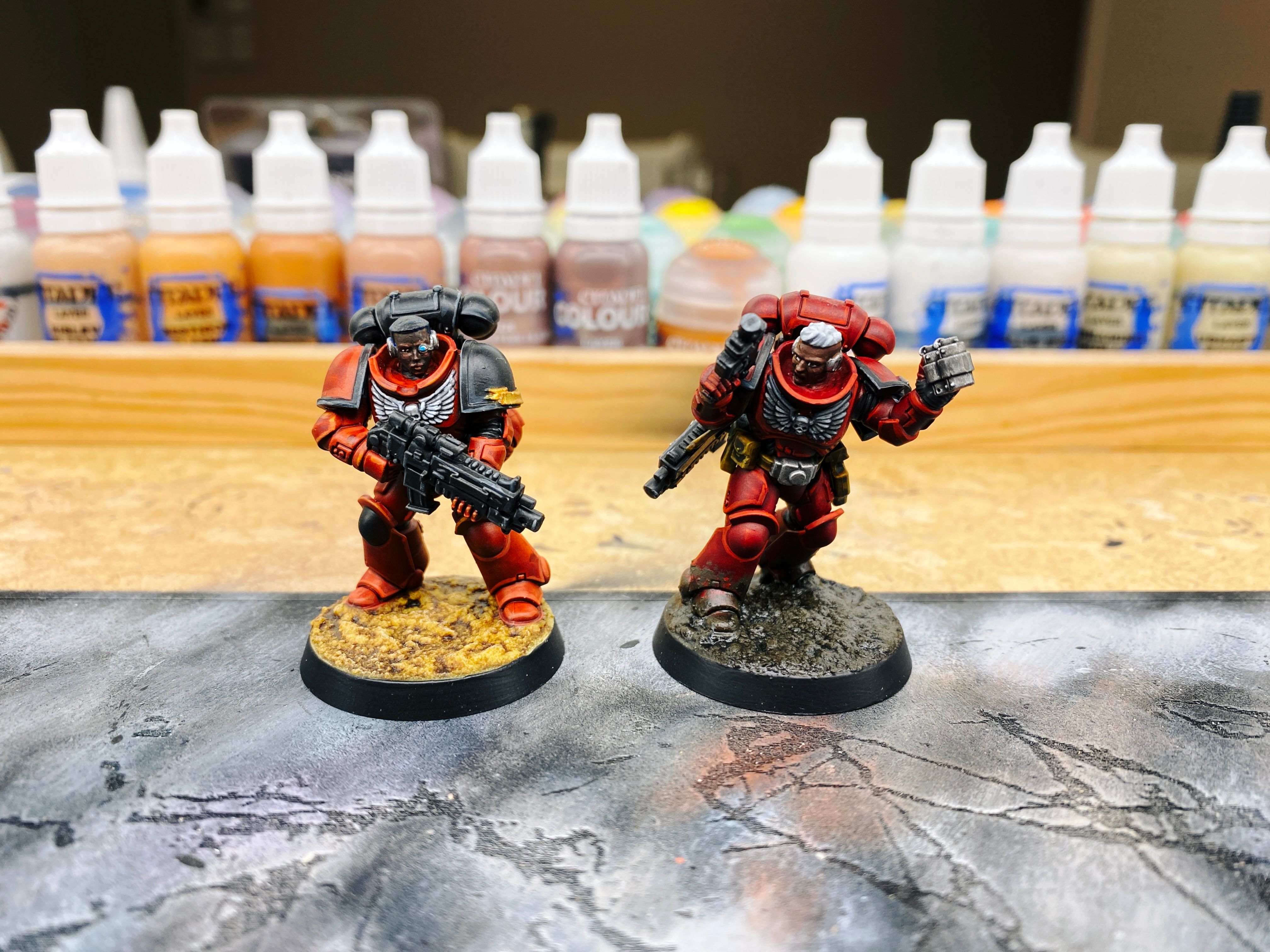 A photo of two fully-painted Space Marine miniatures with bare heads standing next to each other. The left is holding her boltgun in front of like she's standing on guard, and the base is a sort of desert rocky-looking texture/colour. The one on the right is a much deeper richer red, far closer to the colour of blood, and she has a pistol held in one hand and is in the middle of throwing a grenade with the other. Her base looks like really thick mud and it's splattered half-way up to the knees so it looks like she's been really tromping around a battlefield.
