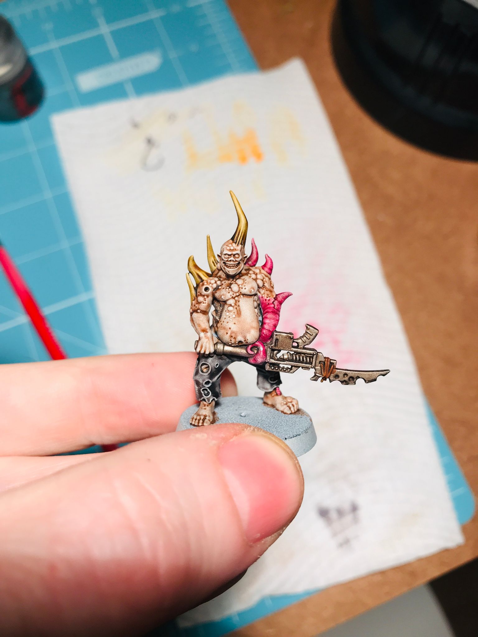 A photo of a Nurgle poxwalker miniature, a fleshy bloated demon with large bone spikes coming out of its head and left shoulder. Its right arm is a pink tentacle, it has the ripped and torn shreds of a pair of dark grey pants, and it's holding a gun  in front of it.