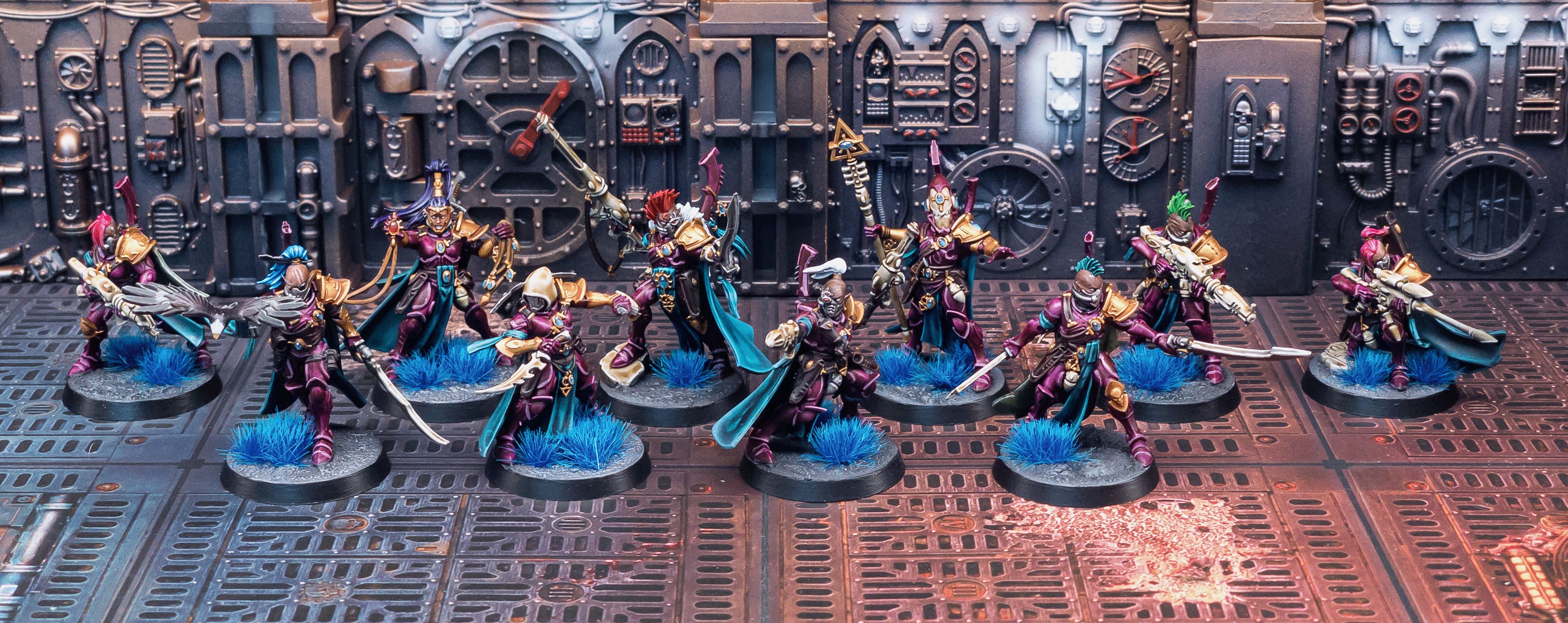 A photo of ten miniatures standing in front of what looks like the inside of an industrial-looking starship. They're essentially space elf pirates, all their armour is elegant and flowing and a rich burgundy colour, and they have cool turquoise capes. They're branding various ranged and close-combat weapons, and several of them have brightly-coloured mohawks.