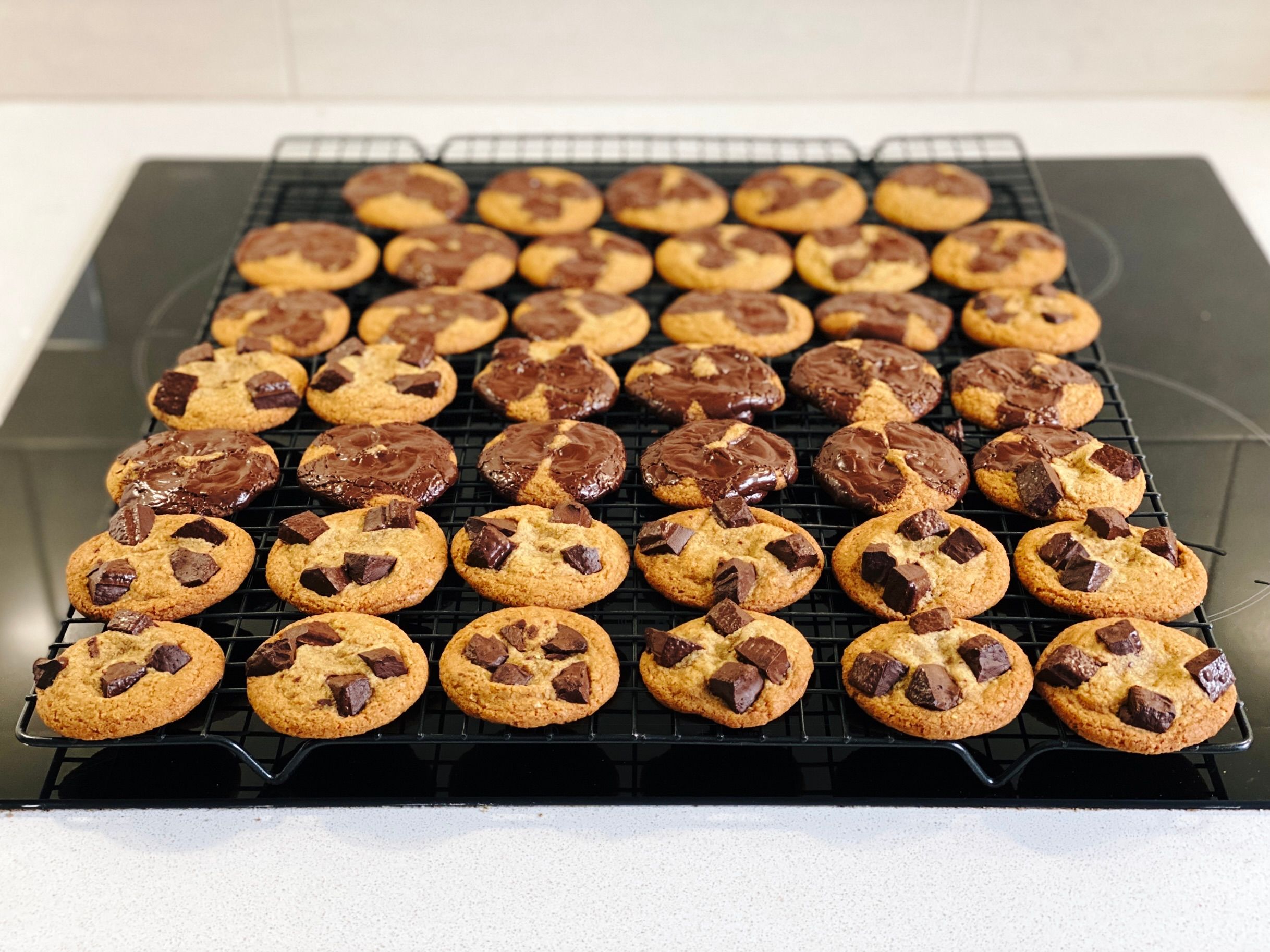 A photo of two cooling racks full of flat, golden-brown cookies. On some of them, there's chunks of chocolate sitting up in the cookie and they look very neat and tidy. On most of the others, the chocolate has totally melted and turned into more of a chocolate coating.