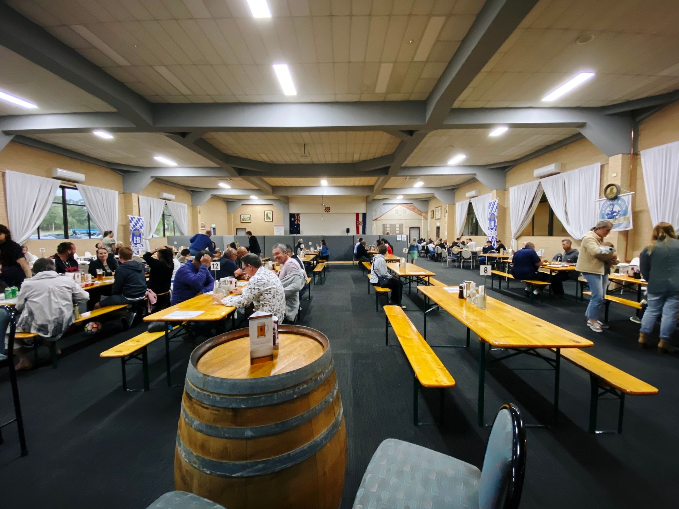 A wide-angle photo of the inside of a big community hall with blue-grey carpet and a bunch a wooden-topped trestle tables, with plenty of people sitting at them.