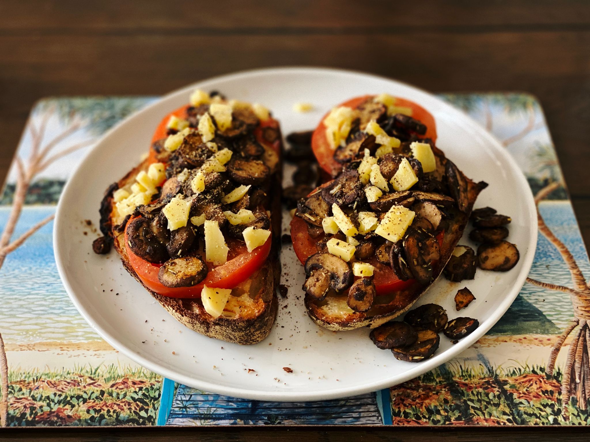 A photo of two slices of homemade bread sitting on a plate, topped with thick slices of tomato with mushrooms on them, and small pieces of crumbled up cheddar cheese atop it all.