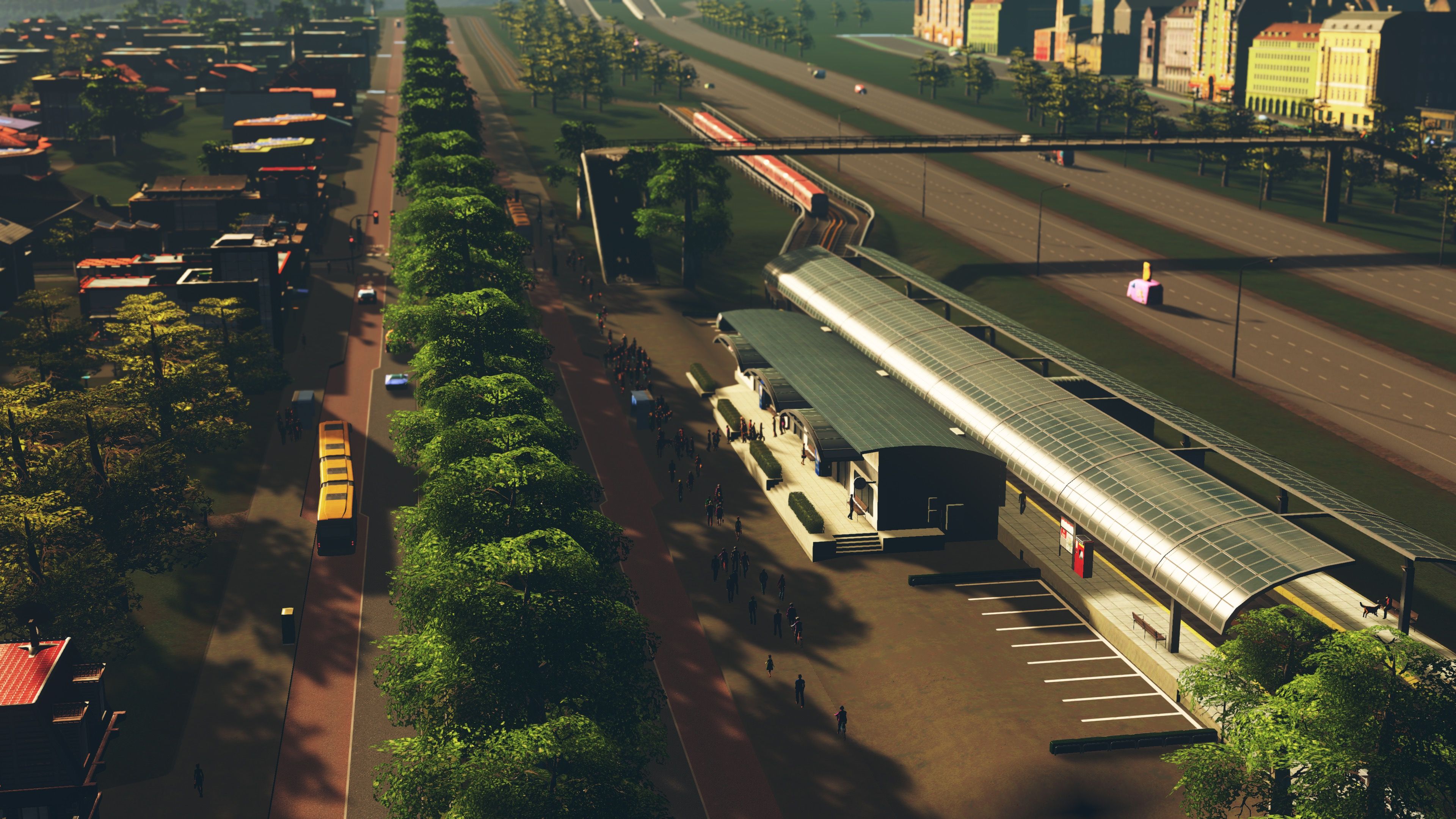 A screenshot from Cities: Skylines, looking down a leafy boulevard. The road has two lanes on each side, one regular and one bus lane, and there's a yellow bendy-bus just pulling into a stop at the left. On the right alongside the road is a train station with a whole bunch of people walking down the street and going into the station. On the other side of the train station is a highway, and there's a pedestrian foot bridge crossing over it.