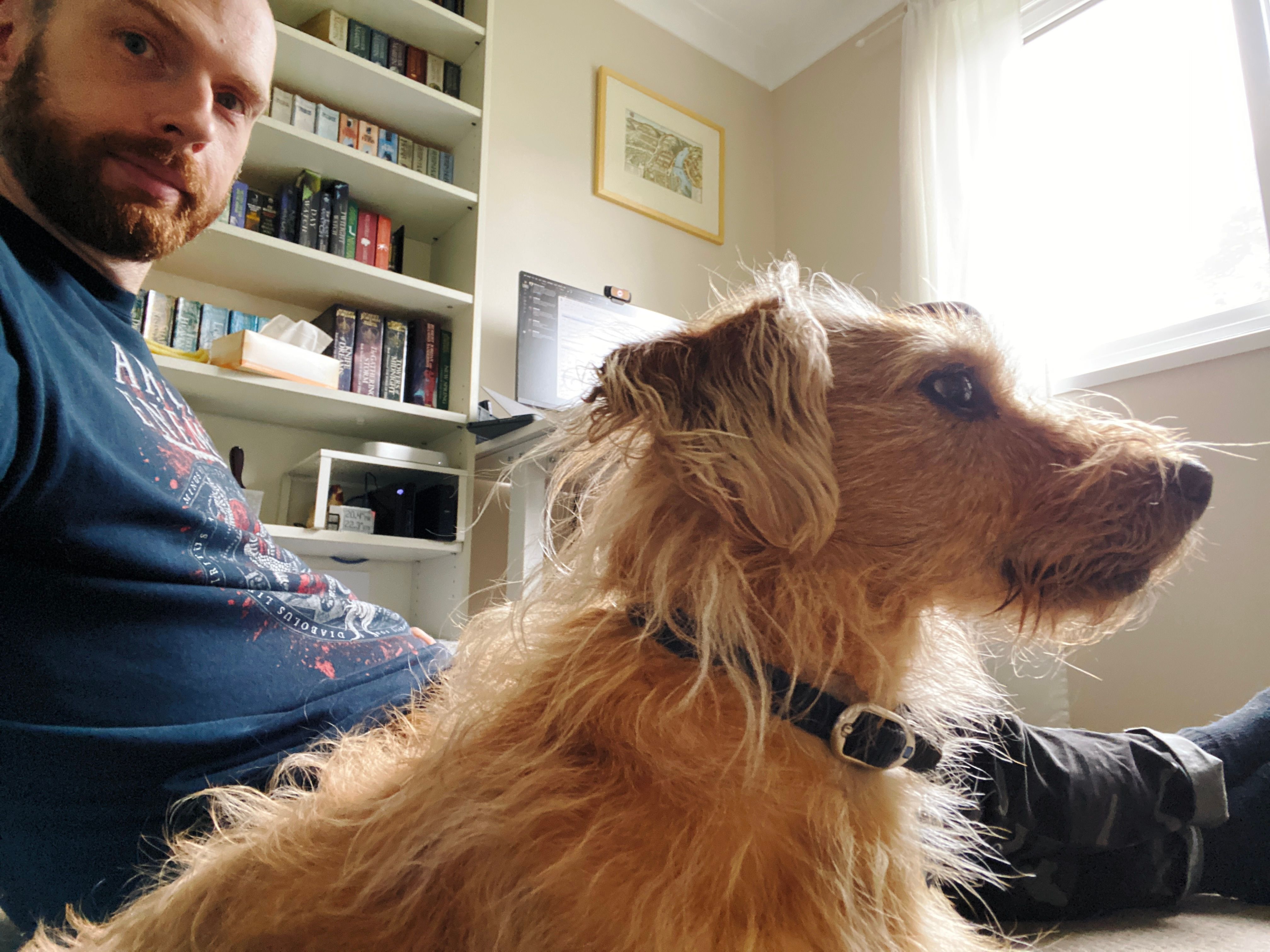 A photo taken from a low angle of the side of a small scruffy blonde dog who's looking off-camera to the right, with me — a white man with a short red beard and a rapidly-receding hairline — sitting on the ground behind him with my legs stretched out.