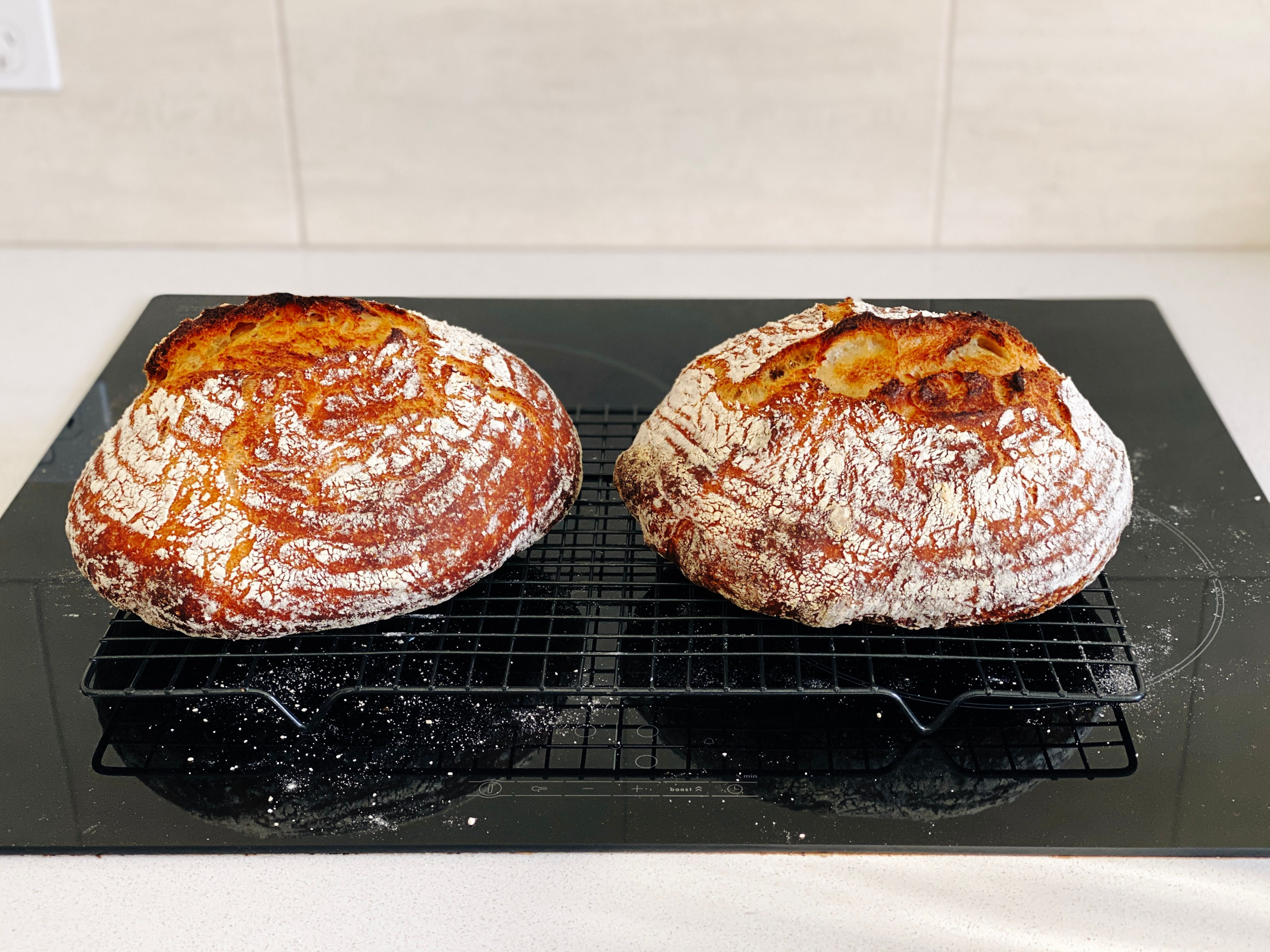 A photo of two golden brown loaves of bread covered in flour, with wonderful-looking splits on top, sitting on a cooling rack.